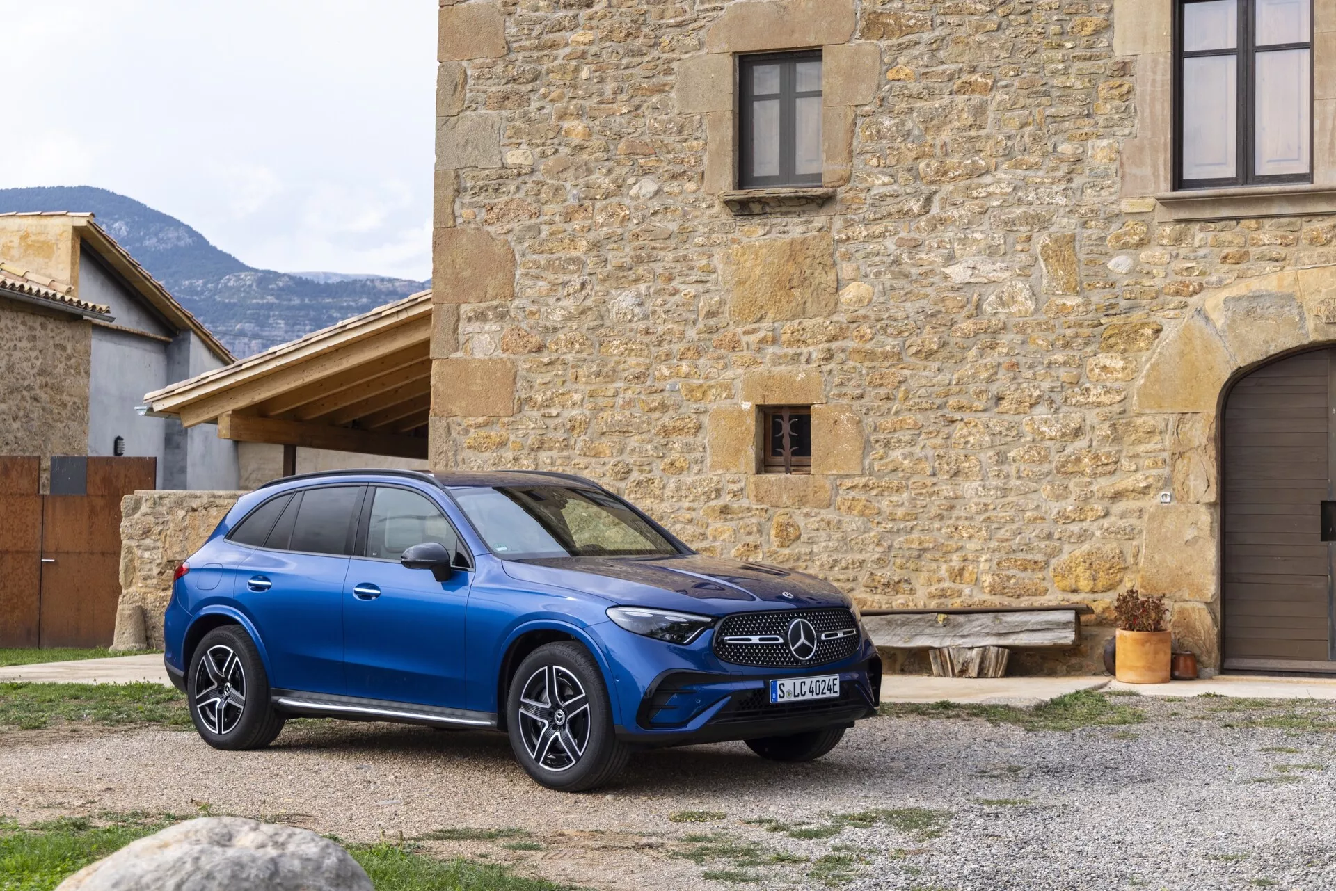 Nowoczesny niebieski Mercedes GLC 2022 SUV jest wyraźnie pokazany na zewnątrz, ustawiony pod lekkim kątem od środka, co podkreśla przedni prawy kwartał oraz część bocznego profilu. Samochód zdobi wyrazisty przedni grill z emblematem producenta, eleganckie poziome reflektory LED oraz duże wieloszprychowe felgi aluminiowe. Jest usytuowany przed starym, kamiennym budynkiem, co zapewnia wyrazisty kontrast. Potencjalne funkcje technologiczne, takie jak adaptacyjny tempomat, asystent pasa ruchu i panoramiczny dach, zostały zasugerowane przez jego design. Kompozycja obrazu oferuje wyraźną, naturalną perspektywę luksusowych i zaawansowanych technologicznie atrybutów pojazdu, ukazując szczegółowy wygląd zewnętrzny w rustykalnym otoczeniu.
