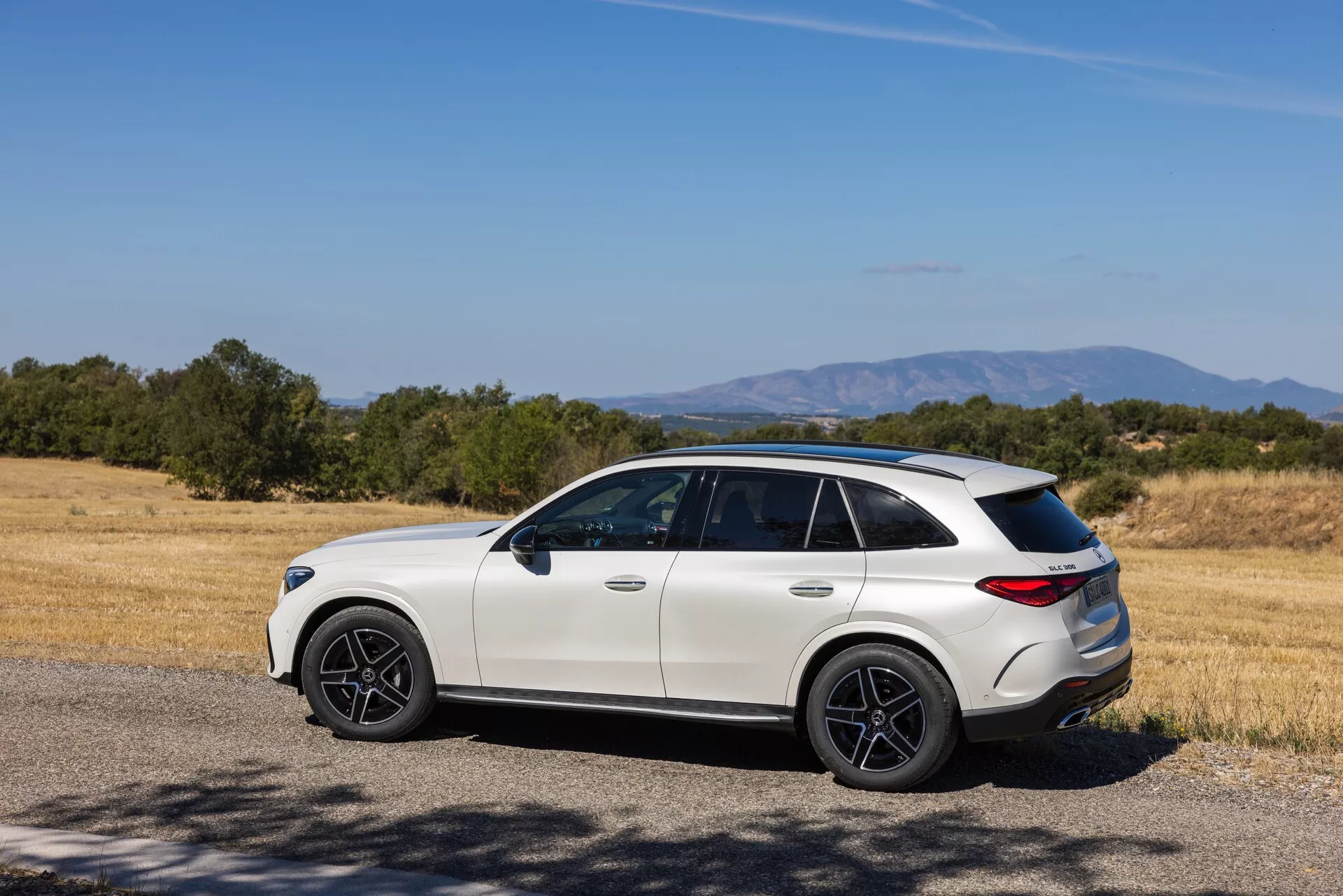 Nowoczesny luksusowy crossover SUV na tle wiejskiego krajobrazu: trzy-czwarte tylno-boczny widok na jasnego Mercedesa GLC 2022 (254) ze sportowymi ciemnymi felgami, przyciemnianymi szybami, pochylonym dachem i rzeźbionym tylnym zderzakiem, zaparkowany w pobliżu suchego pola z trawą i drzewami w pogodny dzień.