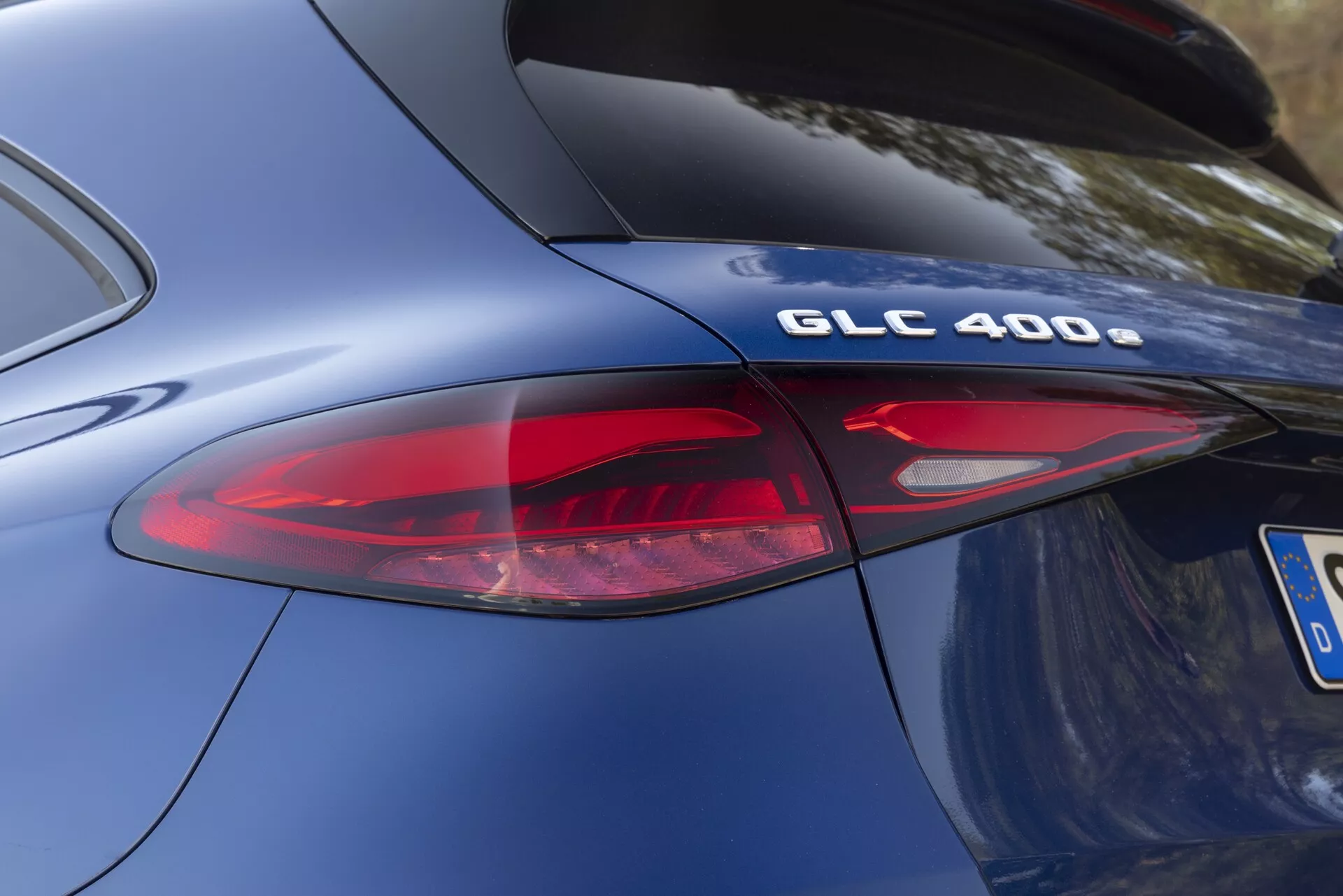 Zbliżony, kątowy widok tylnej części modelu Mercedes-Benz GLC 400 2022. Skupienie na oznaczeniu modelu 'GLC 400' i części nowoczesnego zespołu tylnych świateł LED. Zdjęcie podkreśla krzywiznę karoserii, gładkie metaliczne niebieskie lakierowanie i elegancki design tylnych elementów, w tym czerwonych i przezroczystych elementów soczewkowych świateł tylnych. Chromowane wykończenie na odznace 'GLC 400' sugeruje specyfikacje technologii wydajności i układu napędowego.