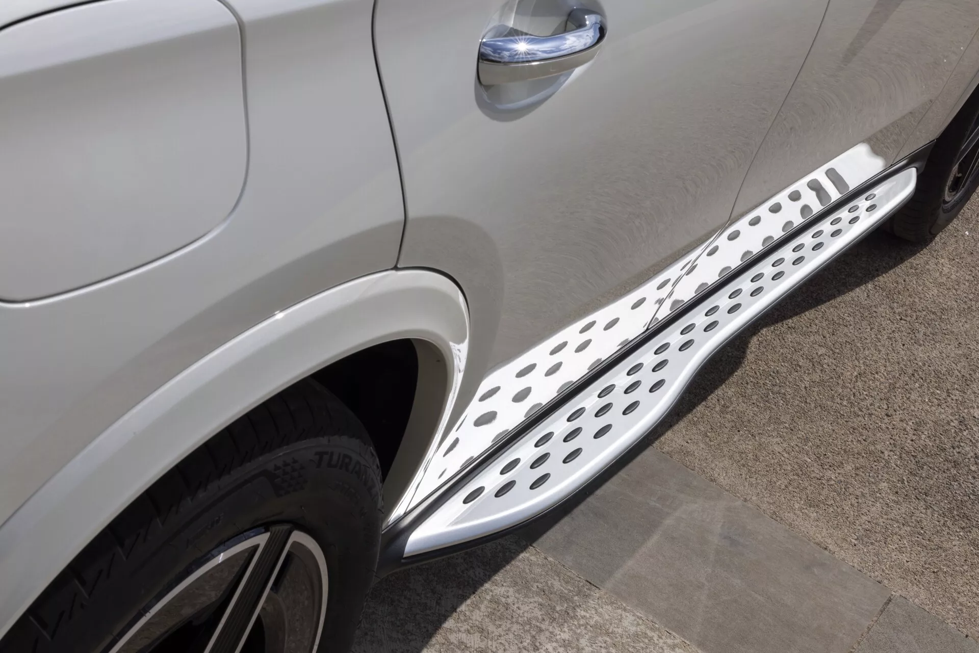 Zbliżenie bocznego widoku Mercedesa GLC 2022 (254) w metalicznym jasno-szarym lub srebrnym kolorze, podkreślające dolną część między przednimi a tylnymi kołami. Widoczna jest część przednich drzwi pasażera i część tylnych drzwi, co wskazuje na model czterodrzwiowy. Metalowy stopień boczny z okrągłymi perforacjami, prawdopodobnie aluminiowy lub ze stali nierdzewnej, biegnie znacznie pomiędzy kołami. Zdjęcie pokazuje również górną część przedniej opony po stronie pasażera z napisem Bridgestone TURANZA. Dodatkowe szczegóły obejmują chromowaną listwę na klamce drzwi i linię formowania na nadwoziu, co wskazuje na luksusowe wykończenie i projekt SUV-a.