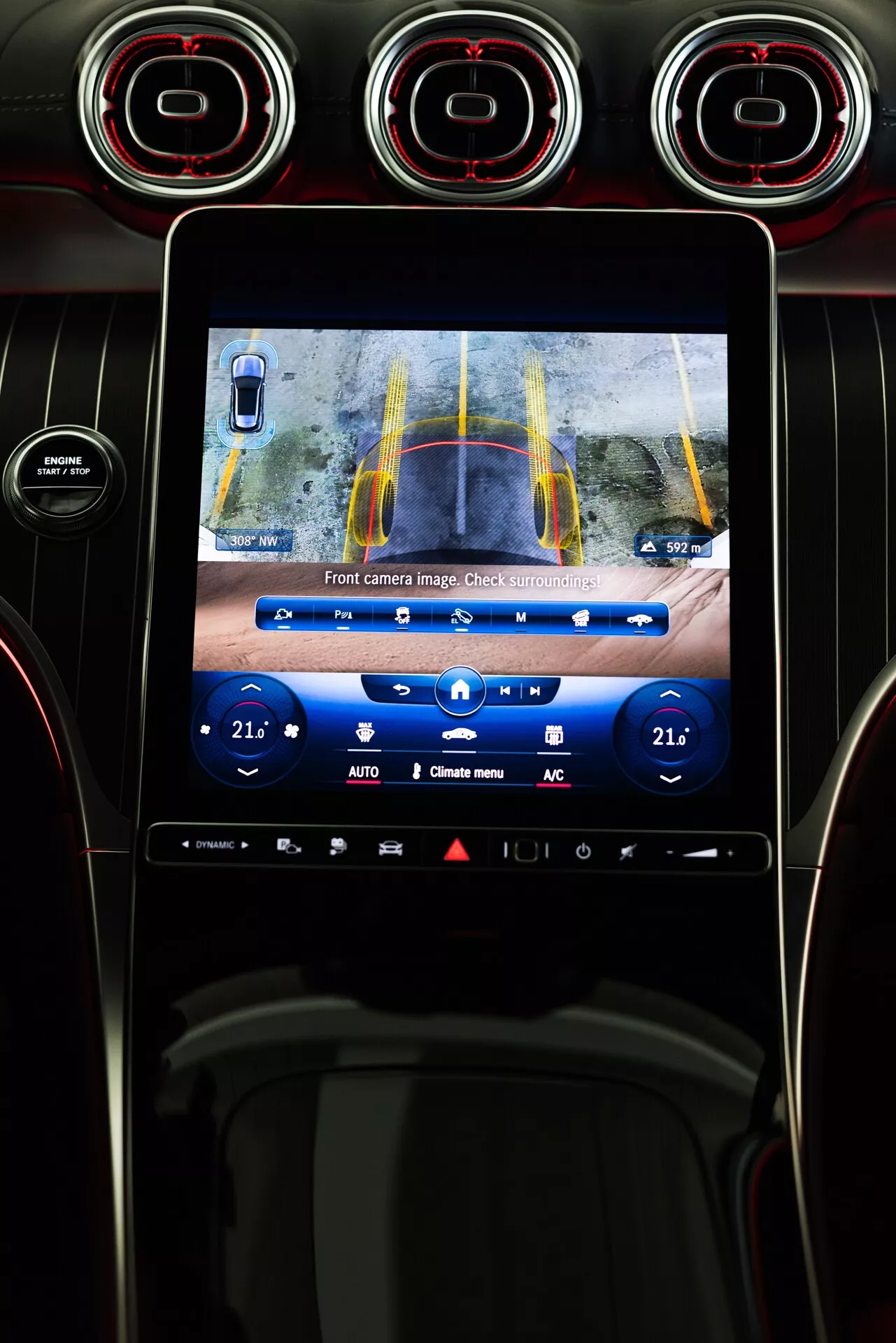 Wnętrze Mercedesa GLC 2022 z centralną konsolą, dotykowym ekranem infotainmentu pokazującym obraz z przedniej kamery z żółtymi i niebieskimi liniami trajektorii do wspomagania parkowania. Widok z fotela kierowcy obejmuje wentylacje z czerwonymi detalami nad ekranem oraz przyciski kontroli klimatu poniżej.