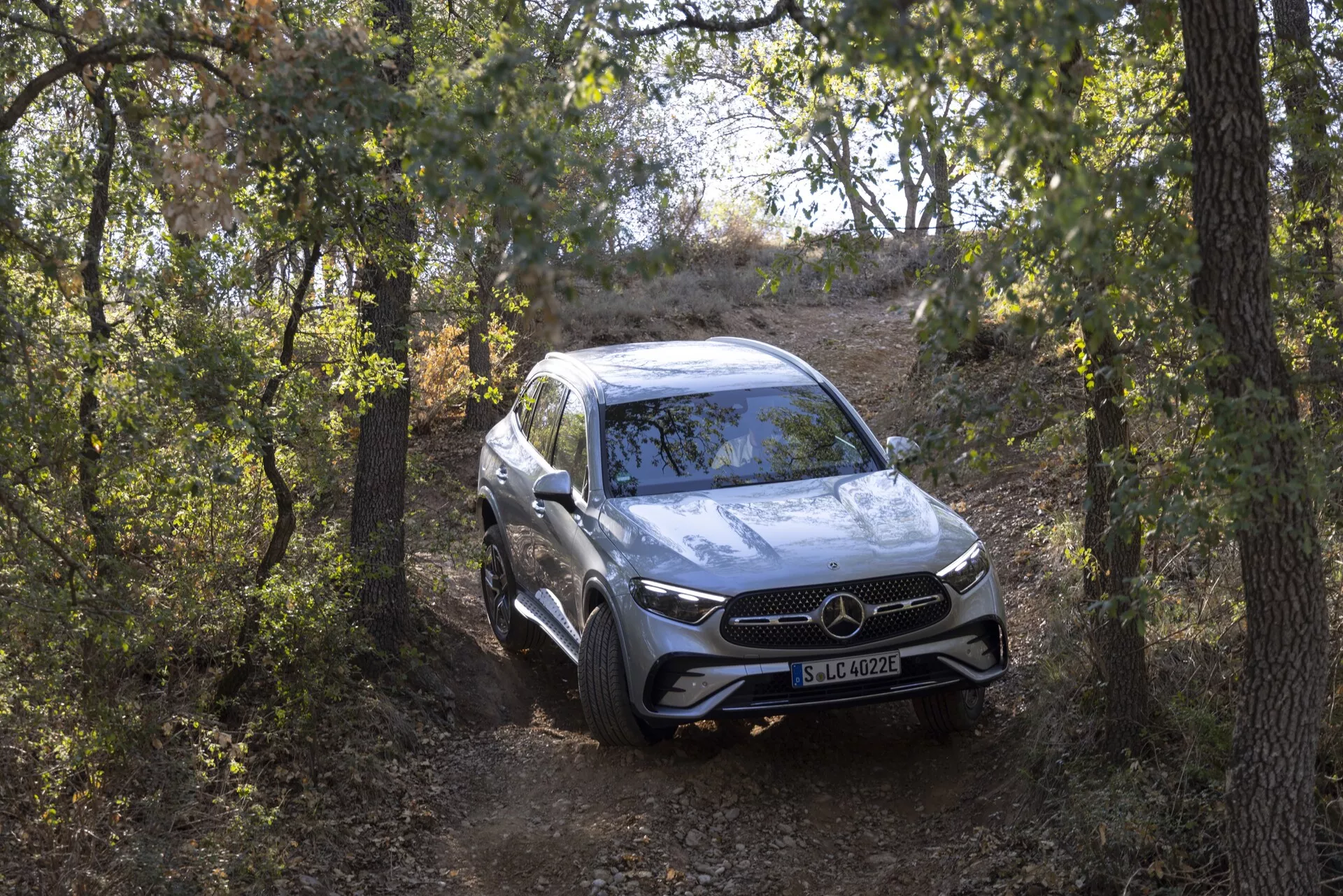 Widok całkowity SUV-a Mercedes GLC 254 z 2022 roku z przedniej perspektywy trzech czwartych, zbliżającego się do kamery na naturalnym szlaku otoczonym drzewami. Lekko wysoki kąt podkreśla jego zdolności terenowe, wysokość jazdy i ustawienie zawieszenia. Pojazd luksusowy posiada wyrazisty przedni grill z dużym emblematem, reflektory LED, czujniki parkowania i aerodynamiczne konturowane linie na masce. Srebrna farba odbija otoczenie, a felgi aluminiowe i wszechstronny bieżnik dodają sportowego i eleganckiego wyglądu.
