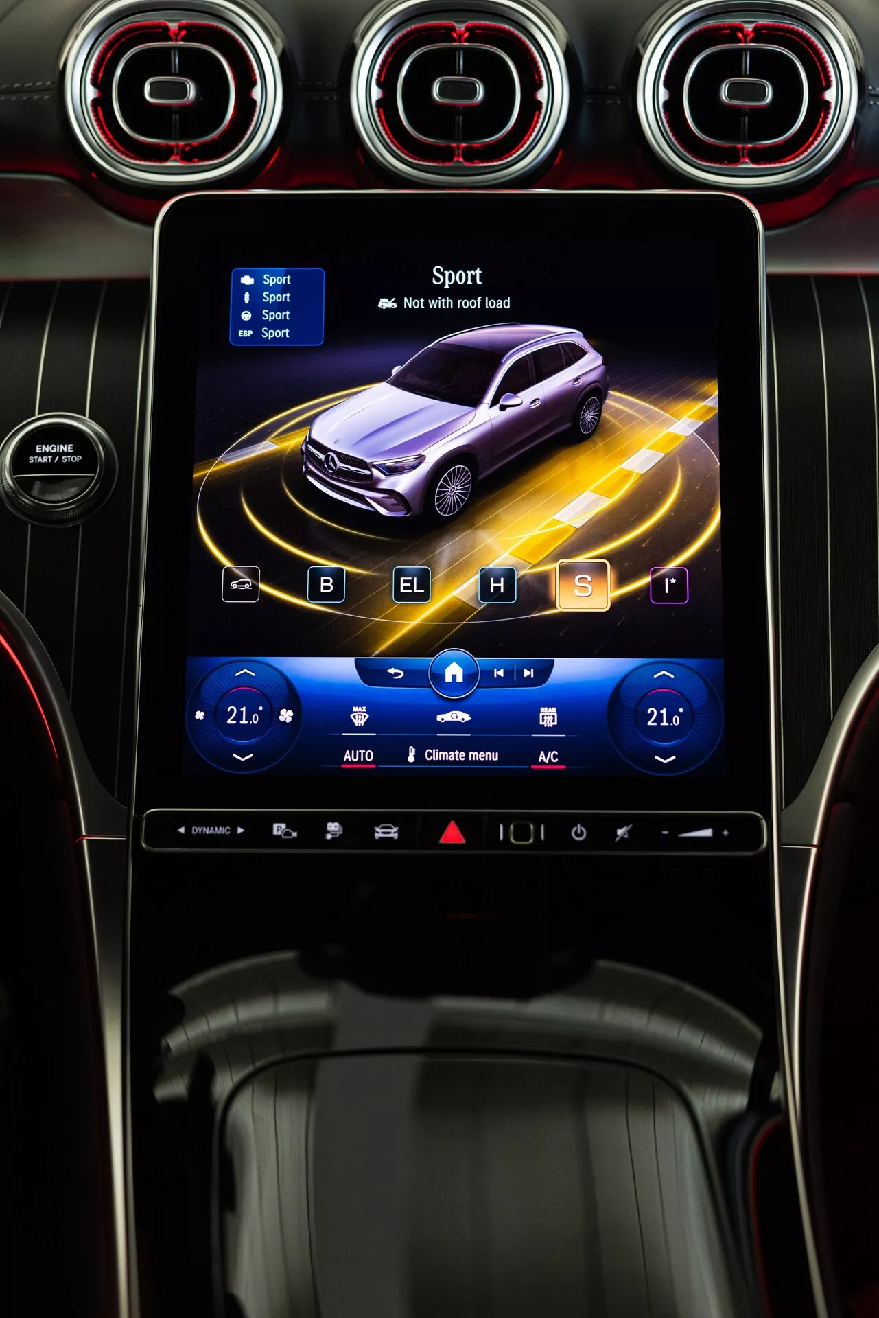 Konsola centralna Mercedesa GLC 2022 z systemem infotainment, pokazującym animowany profil ciemnofioletowego samochodu otoczonego dynamicznymi żółtymi liniami, w trybie 'Sport' z ikoną szachownicy. Ekran dotykowy otoczony jest metalowymi okrągłymi wylotami powietrza z czerwonymi akcentami. Poniżej ekranu, dotykowe sterowanie klimatyzacją pokazuje dwustrefowe temperatury ustawione na 21°C, z ikonami podgrzewania siedzeń, klimatyzacji i odmrażania. Błyszczący czarny panel z dodatkowymi przyciskami dotykowymi poniżej. Styl sugeruje zaawansowaną technologię i sportową estetykę.