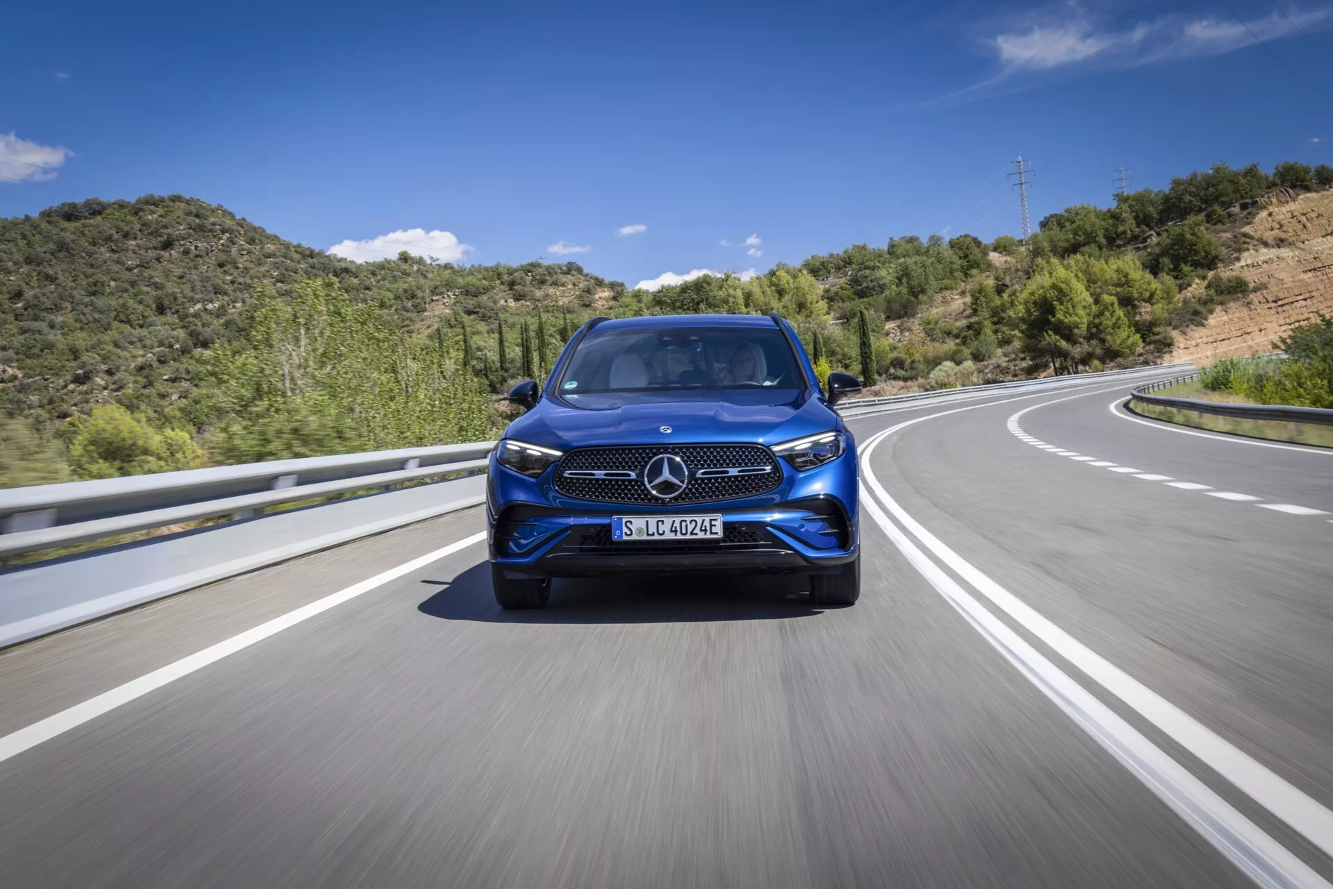 Nisko kątowy, przedni widok nowoczesnego Mercedesa GLC 2022 poruszającego się po autostradzie lub wiejskiej drodze. Ten obraz podkreśla charakterystyczną kratkę pojazdu, projekt reflektorów, stylizację przedniego zderzaka, duże felgi aluminiowe i potencjalne zaawansowane systemy wspomagania kierowcy (ADAS). Oświetlenie jest jasne i naturalne z tłem niebieskiego nieba i zielonych wzgórz, sugerując ruch na asfalcie z oznaczeniami pasa podziału.