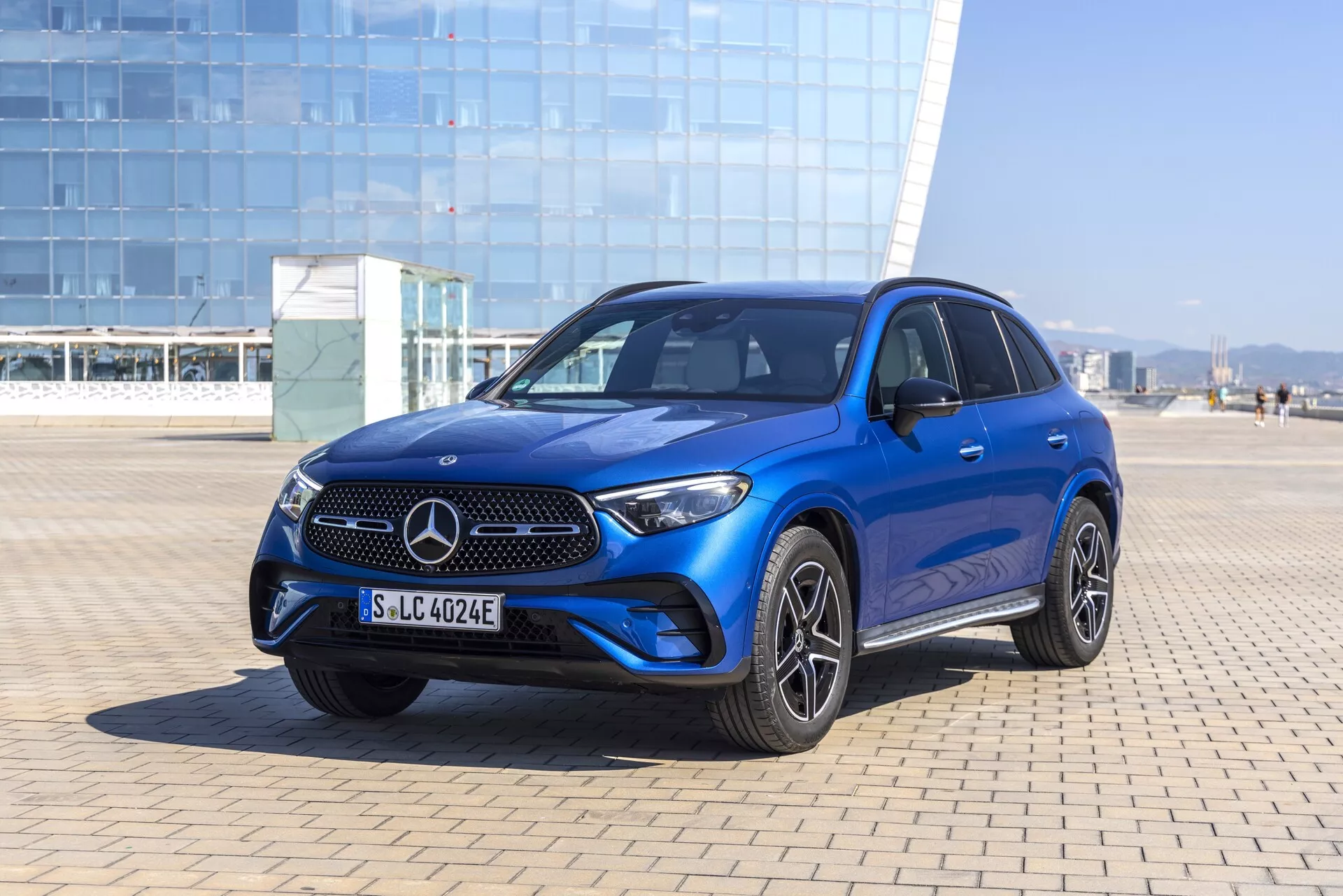 Widok na trzy czwarte z przodu na niebieskiego SUV-a Mercedes-Benz GLC 2022, przedstawiający charakterystyczny grill Mercedes z emblematem, eleganckie poziome listwy, zintegrowany zderzak z lampami przeciwmgielnymi i wlotami powietrza, ostre reflektory LED, konturowane linie maski, sportowe wieloszprychowe felgi aluminiowe, klamki i lusterka w kolorze nadwozia, przyciemniane szyby, szyberdach oraz możliwe zaawansowane systemy wspomagania kierowcy. Zrobione w jasnym plenerze z naturalnym światłem słonecznym i czystym niebem.