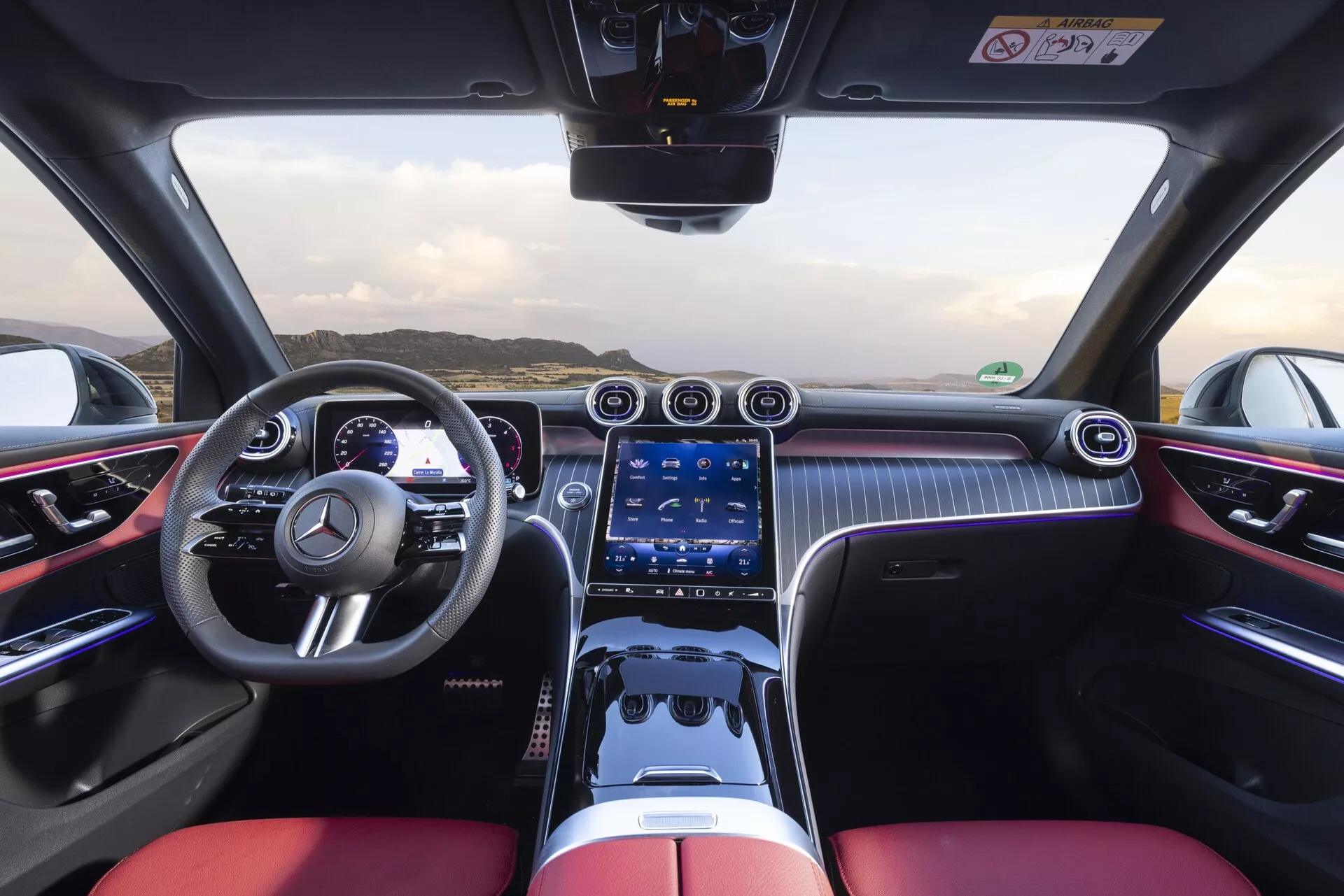 Widok wnętrza Mercedes-Benz GLC 254 z 2022 roku z perspektywy kierowcy, prezentujący sportową kierownicę z wielofunkcyjnymi przyciskami, dwukolorowy kokpit z luksusowymi siedzeniami w kolorze czarnym i jaskrawoczerwonym, cyfrowy zestaw wskaźników, szeroki system multimedialny z ekranem dotykowym, chromowane nawiewy, fizyczne przyciski sterowania klimatyzacją, wzory przypominające włókno węglowe, oświetlenie ambientowe, panel dotykowy i wyrafinowany design zorientowany na wysoką wydajność.