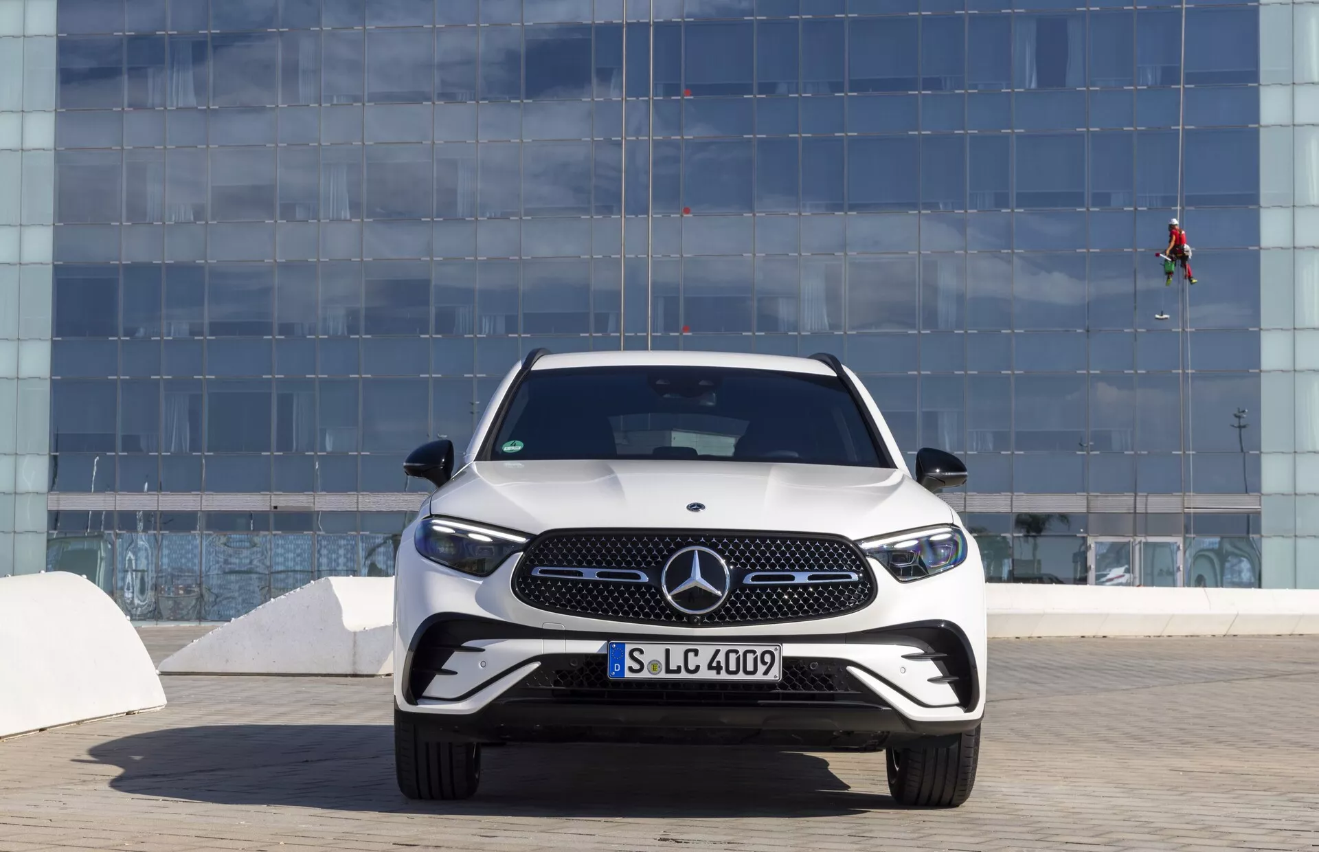 Widok z przodu na Mercedes-Benz GLC 2022 (254) z wyeksponowanym szczegółowym designem przodu, kratką z wyraźnym emblematem Mercedesa, eleganckimi reflektorami LED, sportowym zderzakiem z dużymi wlotami powietrza i aerodynamycznym nagarstem, ustawiony na tle nowoczesnej miejskiej architektury w jasny dzień.