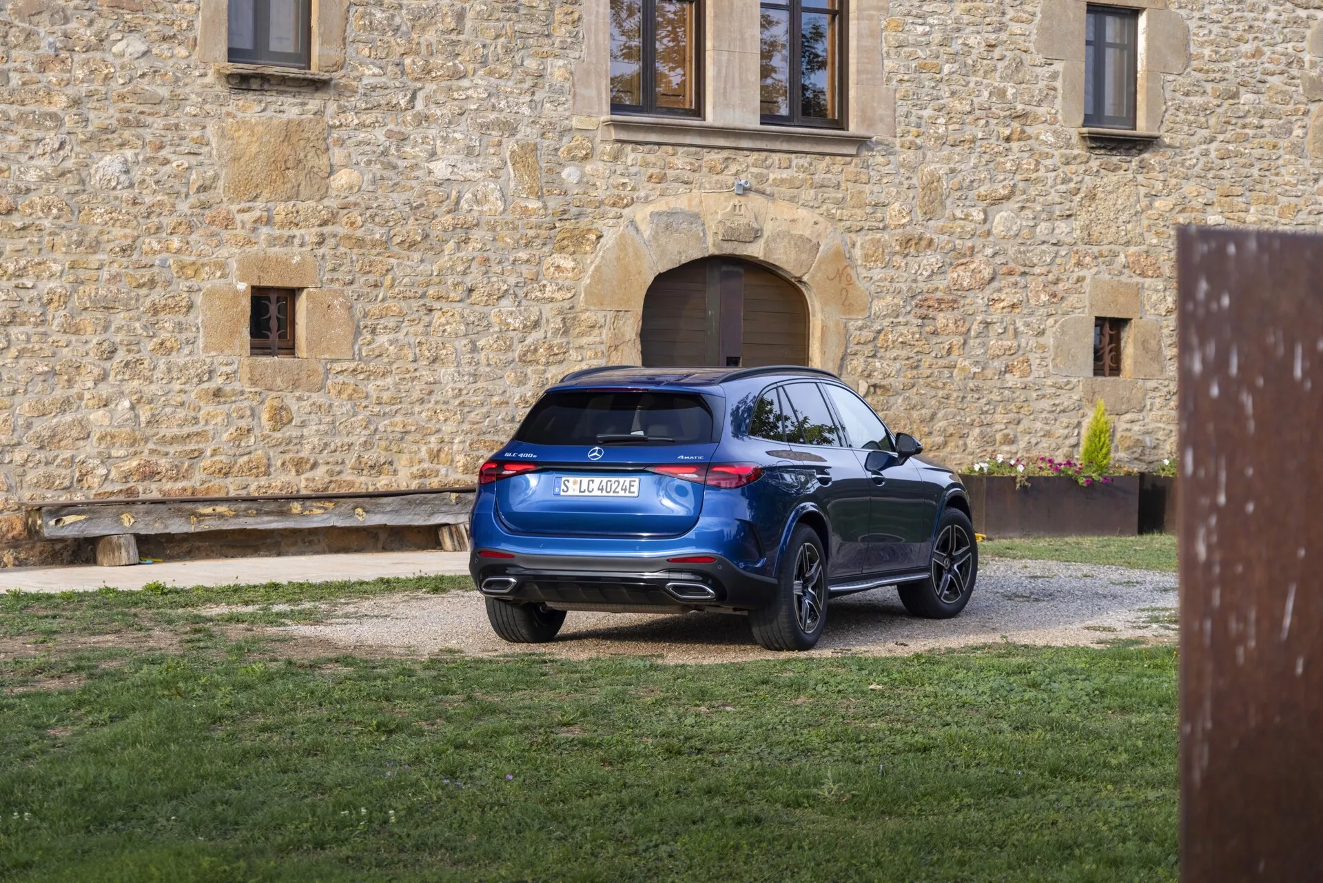 Nowoczesny SUV zaparkowany na żwirowym podjeździe widziany z perspektywy trzech czwartych od tyłu. Tylne światła są wyposażone we współczesną technologię LED, płynnie zintegrowaną z projektem. Lekko nachylona do przodu tylna szyba przyczynia się do aerodynamicznego profilu, uzupełnionego przez spoiler dachowy. Tylny zderzak ma wbudowane czujniki lub reflektory, co sugeruje system wspomagania parkowania, oraz podwójne rury wydechowe w dolnej części zderzaka. SUV ma duże felgi wieloramienne i czystą, bogato niebieską powierzchnię, która odzwierciedla zadbany wygląd na tle rustykalnego budynku z kamienia.