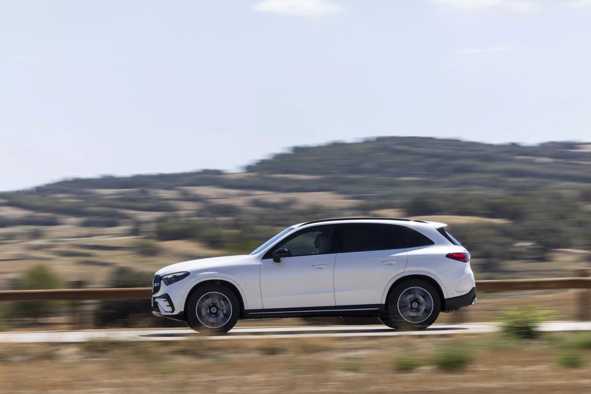 Boczny profil białego SUV-a Mercedes GLC 2022 w ruchu, prezentujący eleganckie linie wzornicze, elegancką karoserię z przyciemnianymi szybami, częściowo rozmyte koła ze stopów, lusterka boczne, wpuszczone klamki drzwi, relingi dachowe i dobrze wyważony kąt kamery z techniką panoramowania, która podkreśla dynamiczny ruch pojazdu.