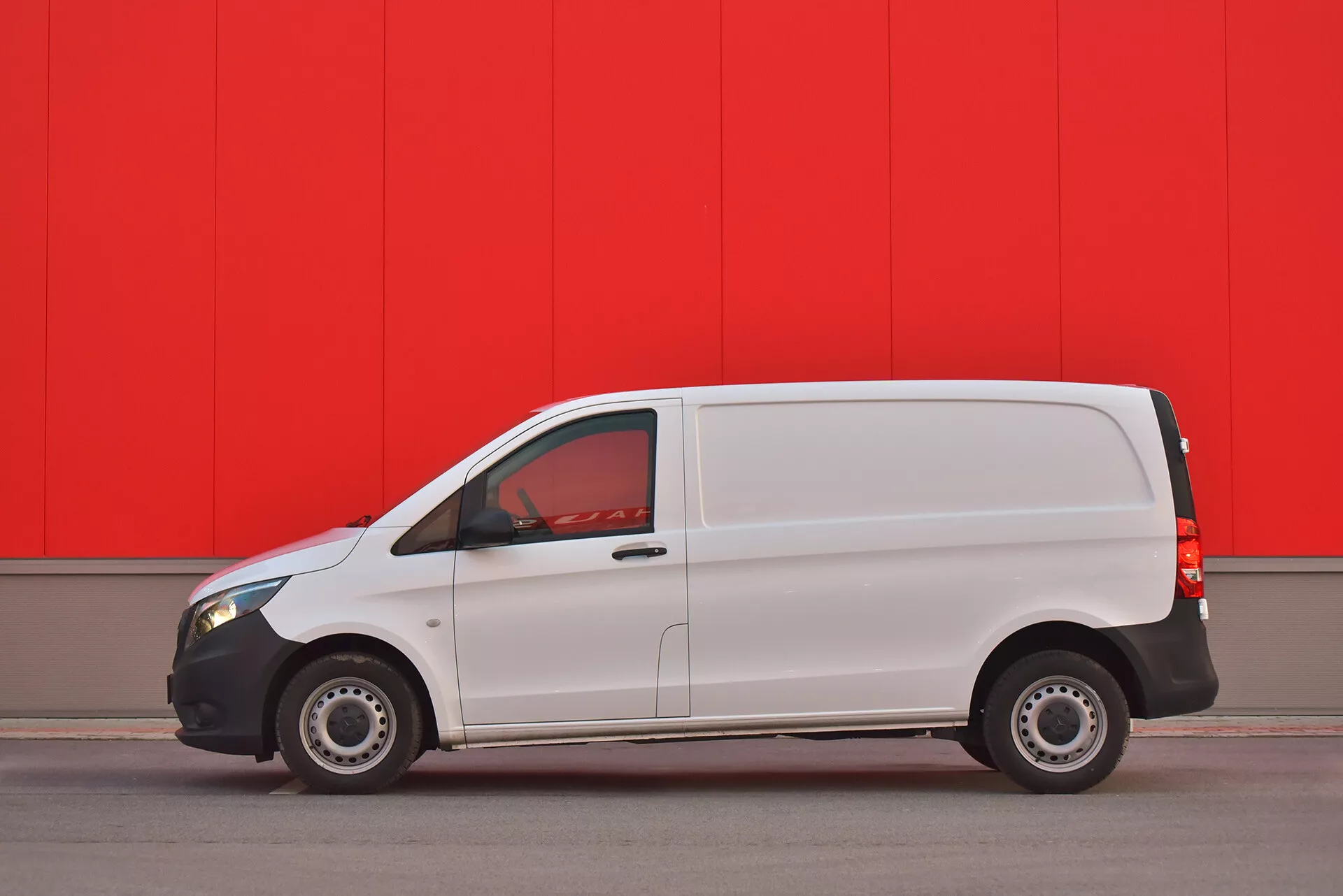 Biały samochód dostawczy Mercedes-Benz Vito Tourer z 2014 roku zaparkowany równolegle przy jaskrawoczerwonej ścianie, ukazujący profil boczny z widocznymi drzwiami, oknami i stalowymi kołami, uchwycony z niskiego kąta, aby podkreślić wygląd i cechy pojazdu