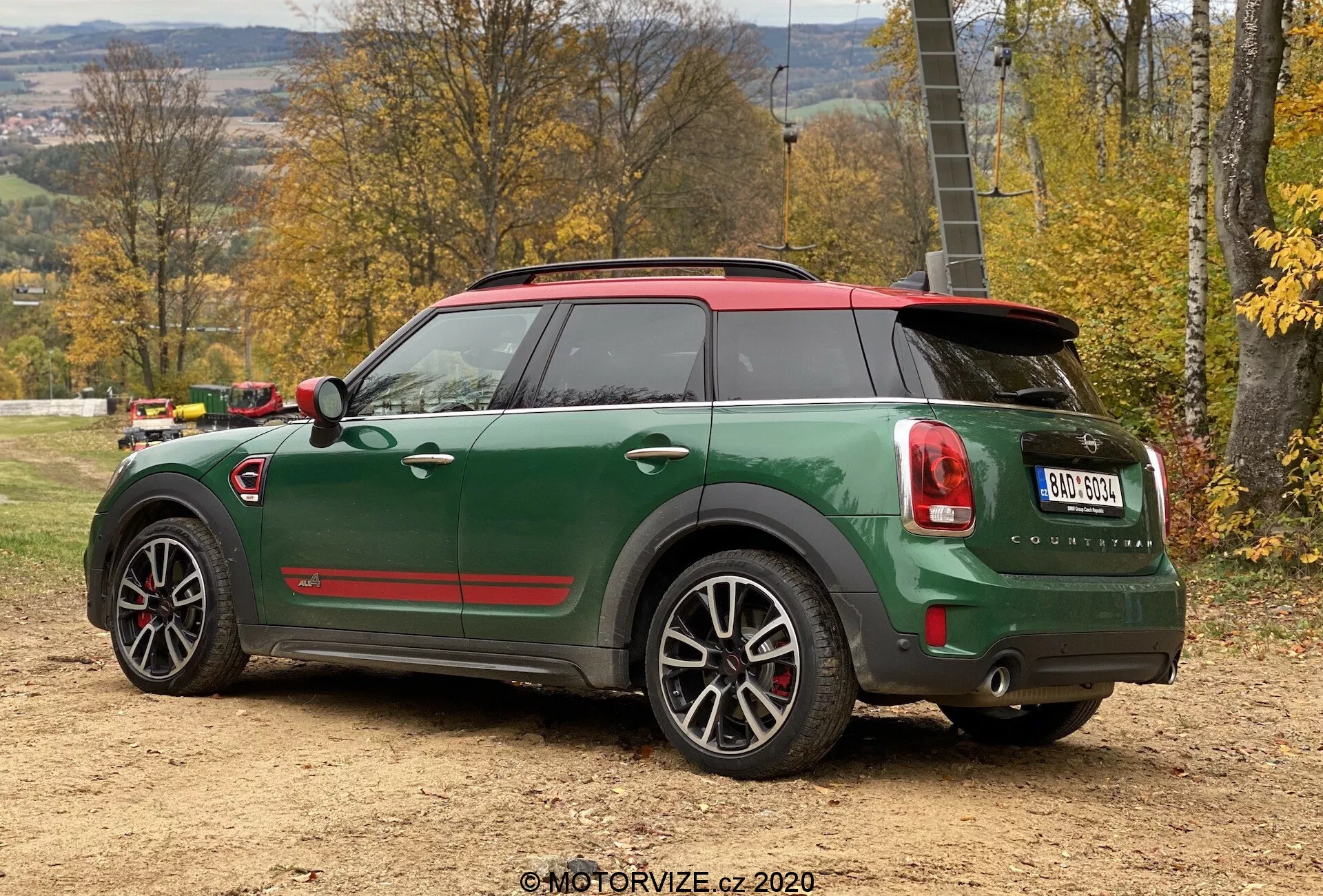 Widok tylno-boczny na zielonego Mini Cooper Countryman z 2020 roku w sportowej wersji John Cooper Works, z czerwonym dachem i osłonami lusterek, podwójnymi końcówkami wydechu, tylnym spojlerem, pięcioramiennymi felgami ze srebrnymi i czarnymi detalami oraz zaawansowanymi funkcjami technologicznymi jak czujniki parkowania i antena typu 'płetwa rekina', ustawionego na nierównej, ziemistej powierzchni w jesiennym, wiejskim otoczeniu.