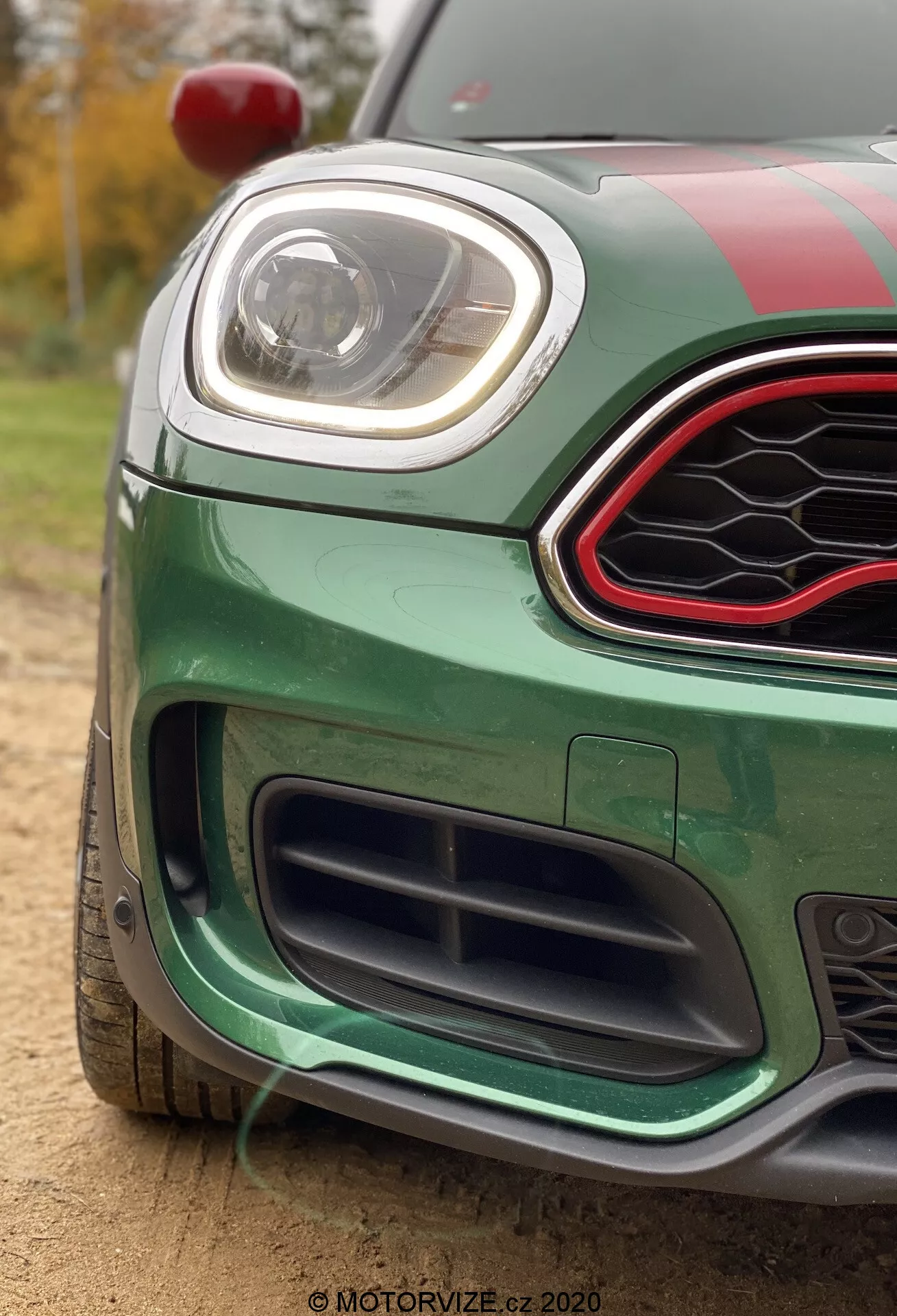 Zbliżenie na zielony Mini Countryman John Cooper Works 2020, koncentrujące się na przednim lewym rogu. Zdjęcie wykonane z niskiego kąta, podkreślające dolną kratkę z czarnym, plastrykowym wzorem, przedni zderzak z aerodynamicznymi elementami i elegancki reflektor LED. Górna kratka ma sześciokątny kształt z błyszczącym czarnym wykończeniem i czerwonym akcentowym obramowaniem. Samochód ma wyścigowe paski na masce i dachu, widoczne czerwone lusterko boczne u góry po lewej, a znajduje się na zewnątrz w naturalnym świetle, z drzewami i roślinnością w rozmytym tle.