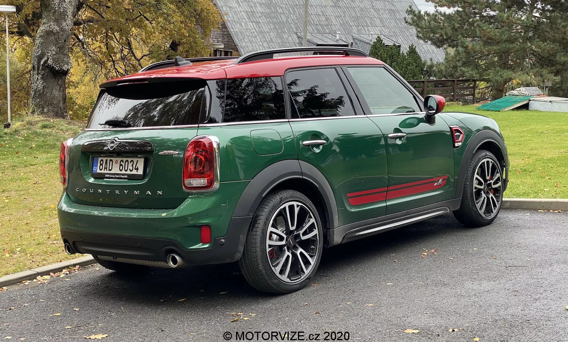 Zdjęcie zielonego 2020 MINI Countryman John Cooper Works w trzech czwartych tyłu, zaparkowanego na podjeździe. Obraz pokazuje całą tylną połowę prawej strony samochodu, w tym tylne drzwi bagażnika, prawe tylne koło, część dachu i drzwi pasażera. Sportowy i kompaktowy design SUV zawiera kontrastujące czerwone akcenty na obudowach bocznych lusterek i dachu, czarne nadkola i duże, wieloramienne felgi aluminiowe. Tylne światła, nazwa modelu 'Countryman' i logo MINI są wyraźnie widoczne. Samochód ma czerwony dach ze spojlerem i relingami, a tylna szyba zawiera odmrażacz i wycieraczkę.