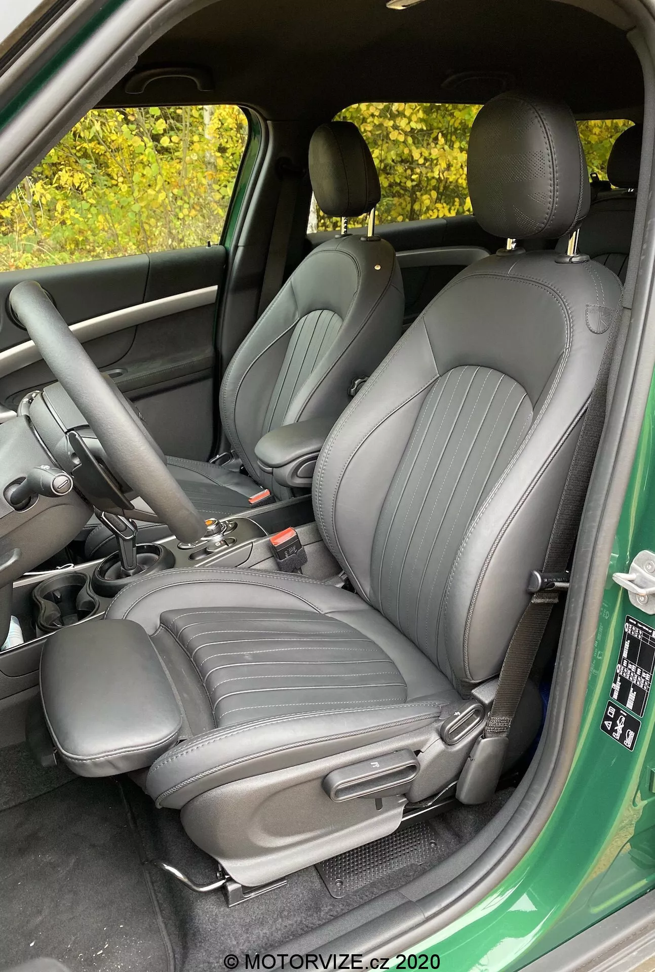 Zdjęcie wnętrza Mini Countryman John Cooper Works 2020 skupiające się na przednich fotelach kierowcy i pasażera, tapicerowanych ciemnym, wysokiej jakości materiałem z pionowymi liniami, widok od otwartych drzwi kierowcy, ukazujący konsolę środkową z dźwignią zmiany biegów i hamulcem ręcznym, częściowy widok panelu drzwi z kontrolkami do okien i lusterek.
