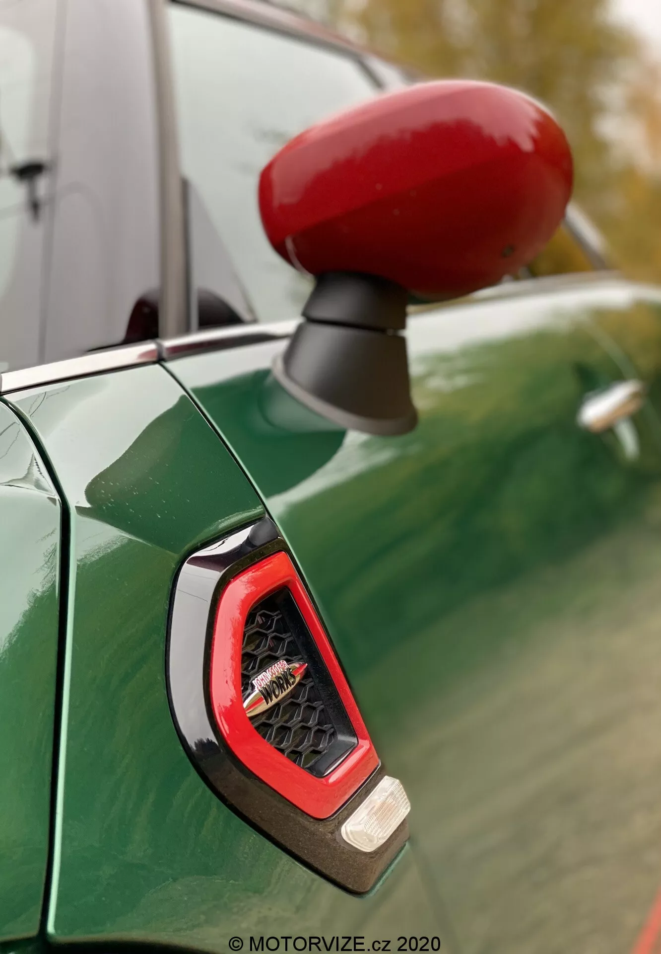 Zbliżenie na intensywnie czerwone obudowę lusterka bocznego i boczny wskaźnik po stronie kierowcy Mini Countryman John Cooper Works 2020, widoczne na tle głębokiego metalicznego zielonego panelu nadwozia z odbiciami pochmurnego nieba i drzew. Skupienie podkreśla kątowy, przejrzysty wskaźnik prawdopodobnie wykorzystujący technologię LED, otoczony sportowym i błyszczącym wykończeniem.