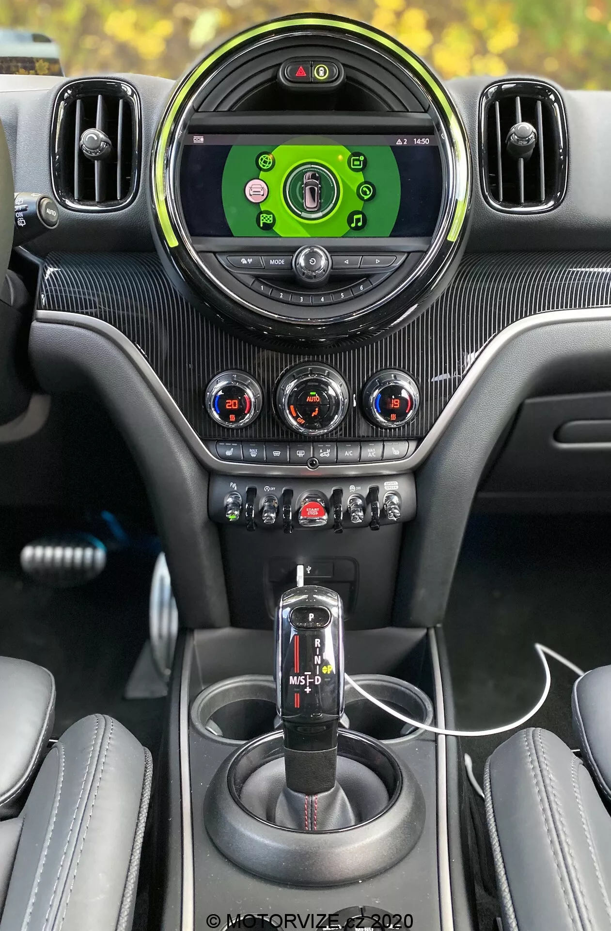 Widok_wnętrza_2020_Mini_Countryman_John_Cooper_Works_z_perspektywy_kierowcy,_podkreślając_premium_funkcje_jak_duży,_okrągły_zegary_zielone_infotaimentu,_otoczone_srebrnymi_regulowanymi_kratek_wentylacyjnych;_trzy_pokrętła_regulacji_klimatyzacji_z_czerwonymi_i_niebieskimi_wskaźnikami_w_panelu_wzoru_włókien_węglowych;_rząd_przycisków_i_przełączników_dla_różnych_funkcji_samochodu,_w_tym_trybu_sport/trakcja;_czarna_dźwignia_automatycznej_skrzyni_biegów_z_opcją_trybu_ręcznego;_spójna_konsola_środkowa_z_wzorem_włókien_węglowych;_dwa_uchwyty_na_kubki;_podłączony_kabel_USB_do_ładowania;_oraz_częściowo_widoczne_skórzane_siedzenia_z_szyciami,_wszystkie_w_pełnej_kombinacji_premium_wewnątrz_samochodu_centrowanego_na_technologiczne-funkcje.