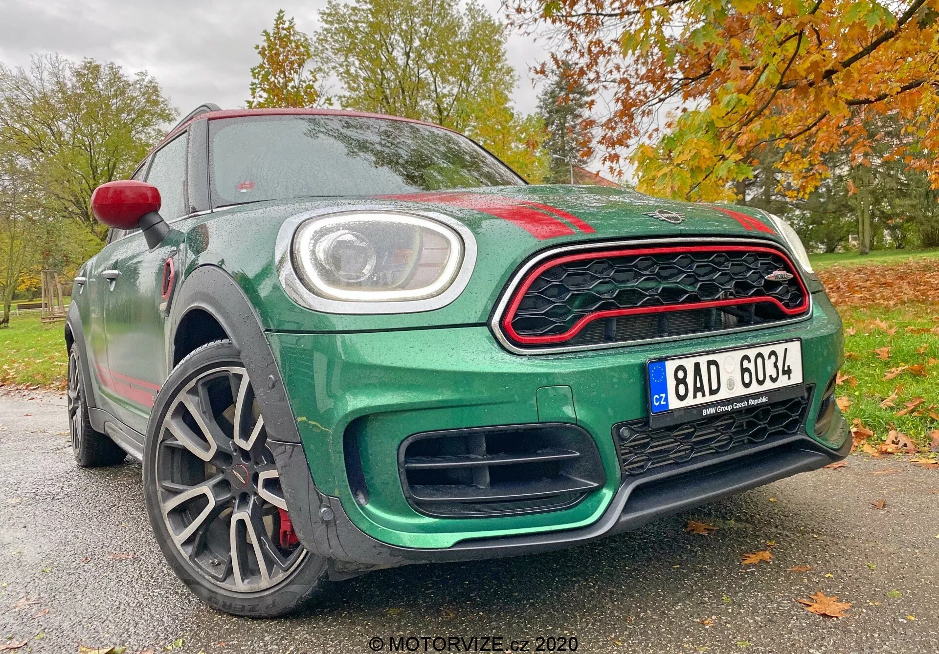 Obraz przedstawia Mini Countryman 2020 edycji John Cooper Works z niskiego kąta przedniego, ukazując jego sportowy i nowoczesny design. Widoczne elementy to unikalny grill przedni w kształcie plastra miodu z czerwonymi akcentami, duże wloty powietrza pod grillem poprawiające przepływ powietrza, centralna tablica rejestracyjna oraz okrągłe światła do jazdy dziennej LED wbudowane w główne reflektory. Maskę wyróżnia lekki wybulenie z aerodynamicznymi konturami, podczas gdy rzeźbione linie bocznego profilu wzmacniają dynamiczny wygląd. Wieloramienne felgi sugerują lekką konstrukcję dla lepszego prowadzenia i przyspieszenia, a także odsłaniają wydajne hamulce tarczowe i zaciski. Karoseria jest pomalowana na bogaty metaliczny zielony, podkreślona przez czerwone lusterko boczne i czerwone akcenty na grillu, na tle jesiennego krajobrazu z liśćmi. Ta perspektywa podkreśla zaawansowane technologicznie i designowe cechy pojazdu, czyniąc go wyróżniającym się autem w kategorii sportowych i luksusowych samochodów.