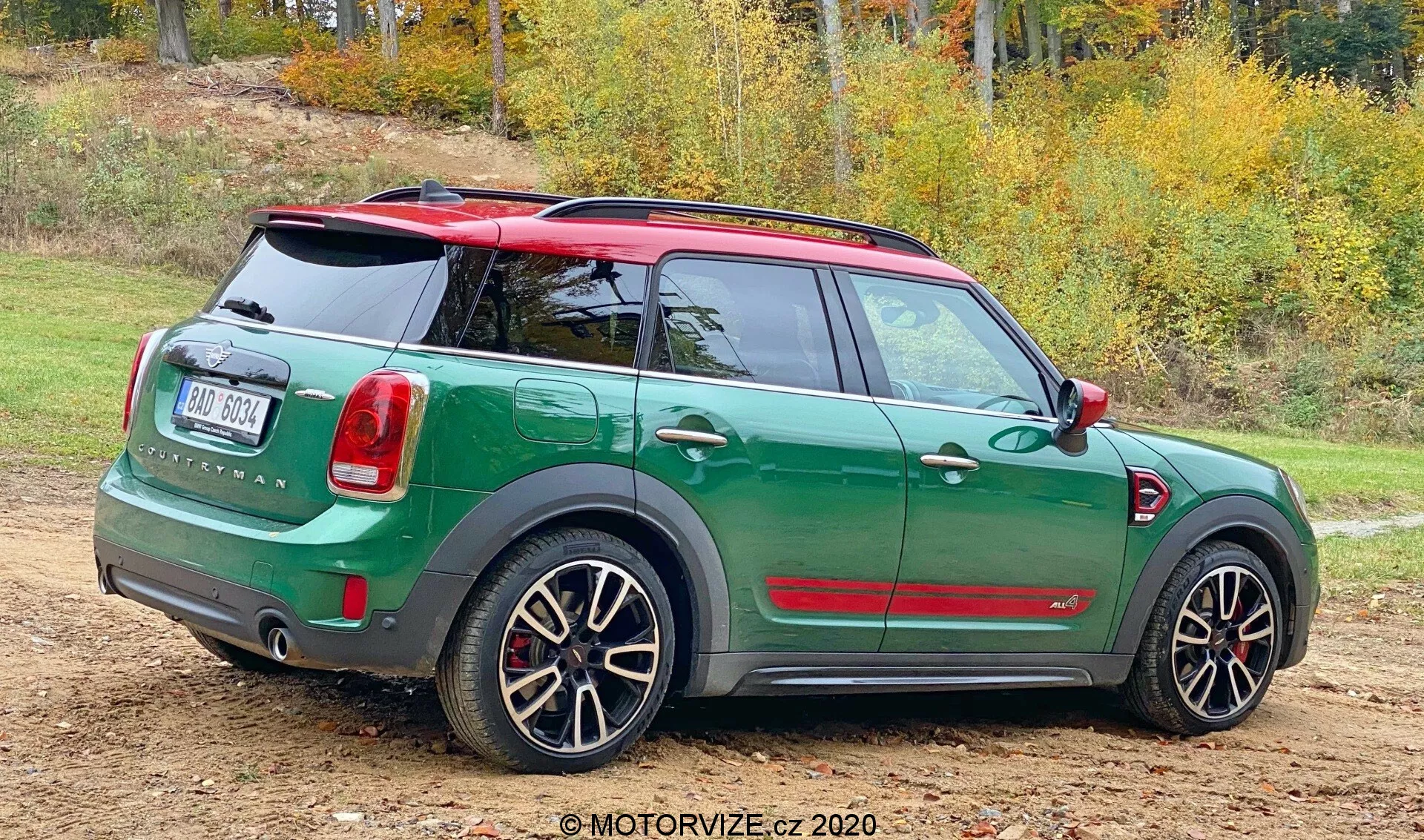 Widok z tyłu (trzy czwarte) Mini Countryman John Cooper Works 2020, zaparkowanego na nieutwardzonej powierzchni z łatami trawy. Sportowe, kompaktowe auto ma dwukolorowe wykończenie z ciemnozielonym nadwoziem i kontrastowym czerwonym dachem oraz lusterkami. Tył samochodu ukazuje tylne drzwi, wycieraczkę, widoczne logo oraz pionowo ustawione tylne światła. Tylny zderzak zawiera czujniki parkowania. Samochód posiada także sportowe, wieloramienne felgi, przez które widać zaciski hamulcowe, co sugeruje wersję o wyższych osiągach. Naturalne tło z jesiennymi drzewami sugeruje porę roku.