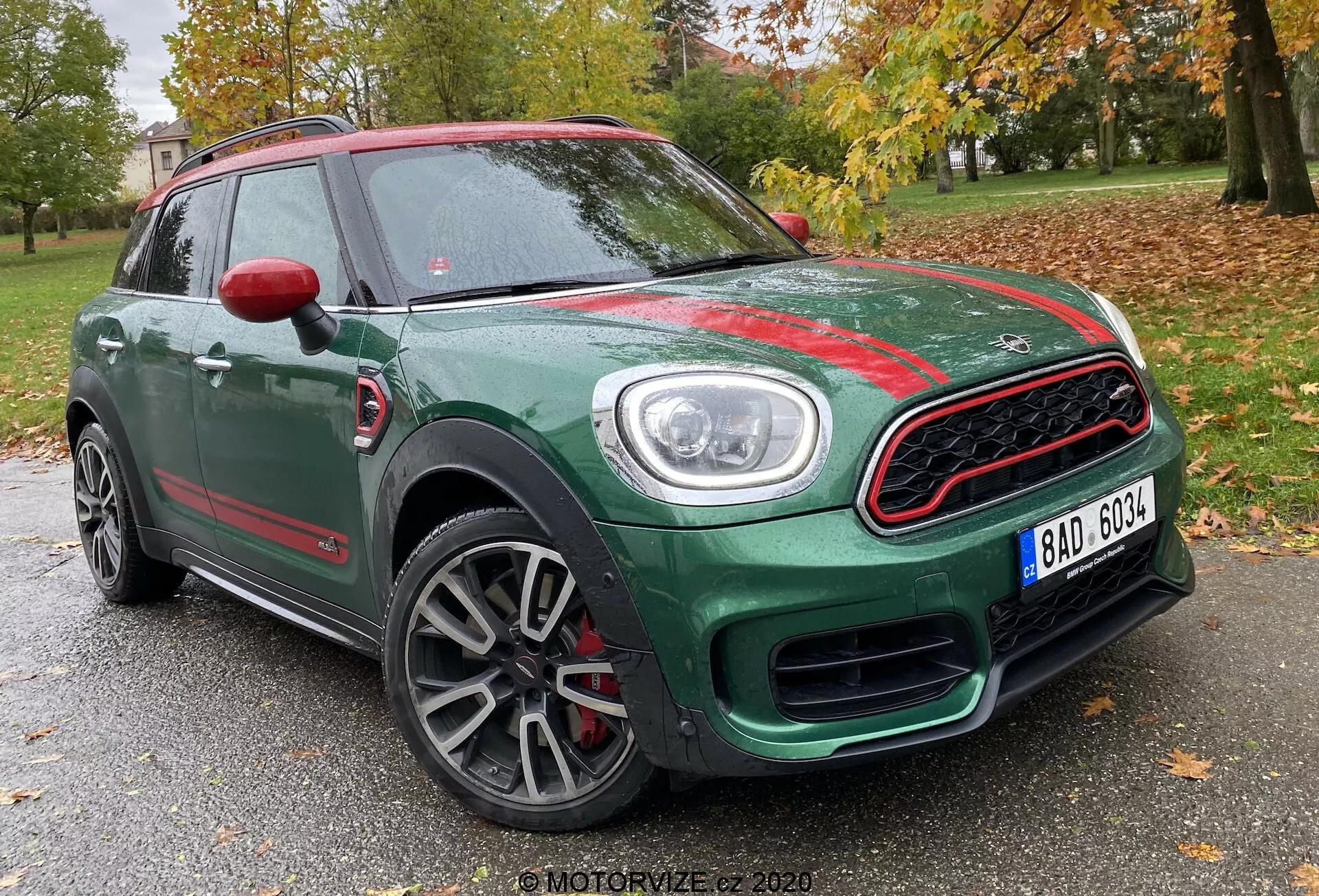 Widok z lewej trzech czwartych przodu Mini Countryman John Cooper Works z 2020 roku, ukazujący jego zielone nadwozie kompaktowego SUV-a z kontrastującymi czerwonymi akcentami. Uchwycone z niskiego kąta, zdjęcie uwypukla sportową przednią osłonę z chromowanym obramowaniem i wzorem plastra miodu, wyraźne okrągłe reflektory z dziennymi światłami LED, wielopromienne aluminiowe obręcze kół z szarym wykończeniem oraz czerwone kapturki bocznych lusterek. Zdjęcie wykonano na tle drzew i słabej struktury, co sugeruje lokalizację w parku lub obszarze mieszkalnym.