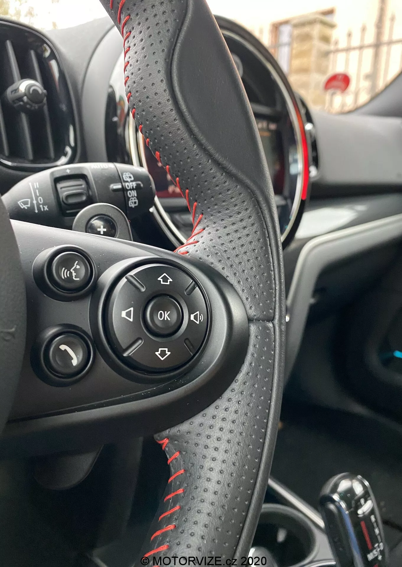 Widok wnętrza Mini Countryman John Cooper Works 2020 skoncentrowany na kierownicy obszytej skórą z czerwonymi przeszyciami, przyciskach sterowania dźwiękiem i połączeniami oraz częściowo widocznym zestawie wskaźników z czerwonymi akcentami, wskazującymi na sportowy model. Pokazuje panel sterowania kierowcy, automatyczną dźwignię zmiany biegów i przyciski sterowania oknami, podkreślając wygodę i zaawansowane funkcje.