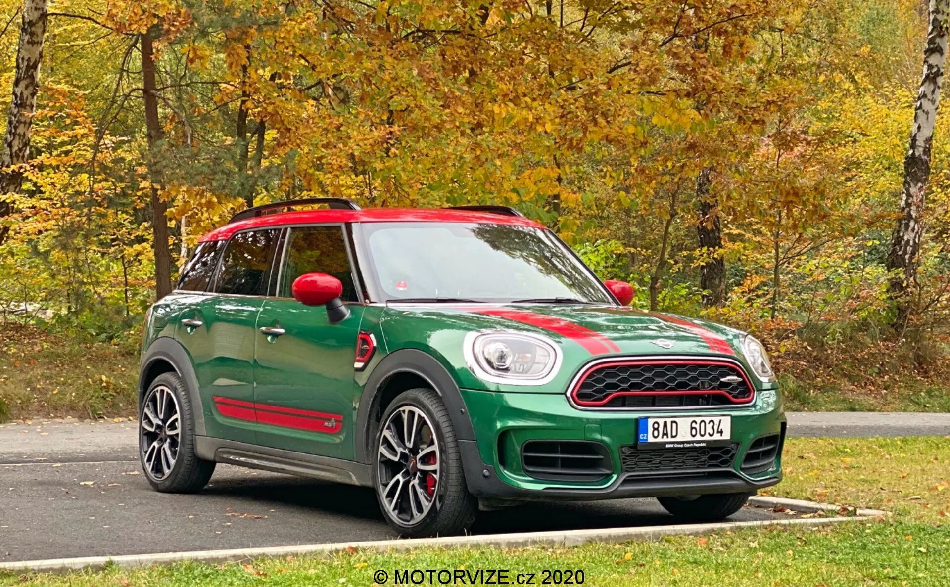 Zielony Mini Countryman John Cooper Works 2020 uchwycony z widoku od frontu z boku. Fotografia ukazuje zarówno przód, jak i bok samochodu, podkreślając jego cechy projektowe. Przód ma charakterystyczny grill z tablicą rejestracyjną poniżej i klasyczne okrągłe reflektory. Sportowe pasy biegną wzdłuż maski, a profil boczny pokazuje felgi ze stopów metali lekkich, lusterka boczne i linie dachu. Czerwone lusterka boczne i pasujący dach tworzą dwukolorowy schemat kolorów. Pod drzwiami, dopasowane progi boczne sugerują zaktualizowany poziom wyposażenia lub pakiet sportowy. Pojazd ma panoramiczny dach i jest zaparkowany na nawierzchni asfaltowej z jesiennym, naturalnym tłem. Ostre, jasne zdjęcie wykonano w naturalnym świetle dziennym.