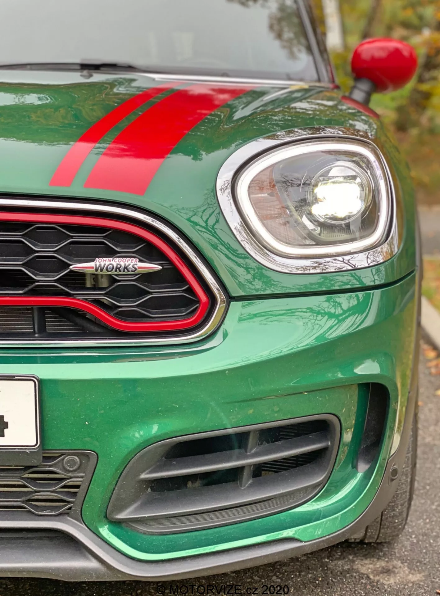 Zbliżenie na przedni lewy narożnik 2020 Mini Countryman John Cooper Works. Obraz, wykonany nieco poniżej poziomu reflektorów, podkreśla metaliczny zielony maskę z czerwoną wyścigową paskiem, okrągły lewy reflektor otoczony chromem i część kratki w kształcie plastra miodu z błyszczącym czarnym otoczeniem. Widoczne są odznaka 'John Cooper Works', lewy wskaźnik na nadkolu oraz duże wloty powietrza w przednim zderzaku zaprojektowane do chłodzenia silnika i aerodynamiki. Samochód wygląda na czysty i dobrze utrzymany, podkreślając nowoczesną technologię reflektorów z projektorem lub LED oraz sportowy, ukierunkowany na osiągi design.