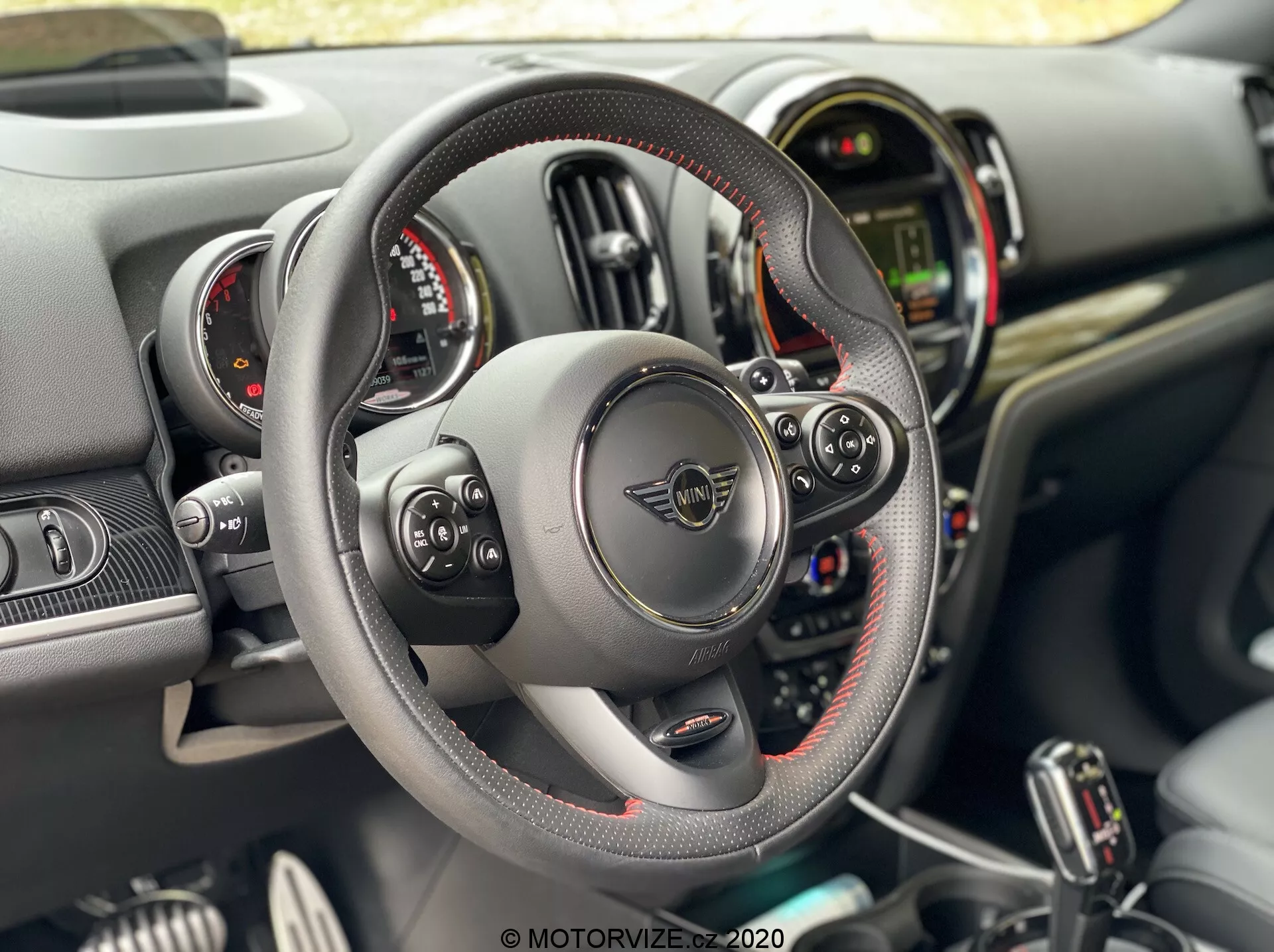 Wnętrze Mini Countryman John Cooper Works 2020 z perspektywy kierowcy, z kierownicą z płaskim dnem obszytą skórą i widocznymi szwami oraz wielofunkcyjnymi przyciskami, łopatkami zmiany biegów za kierownicą, podwójnymi okrągłymi tarczami instrumentów otaczającymi centralny wyświetlacz cyfrowy oraz deską rozdzielczą z różnymi przełącznikami i przyciskami na konsoli centralnej.