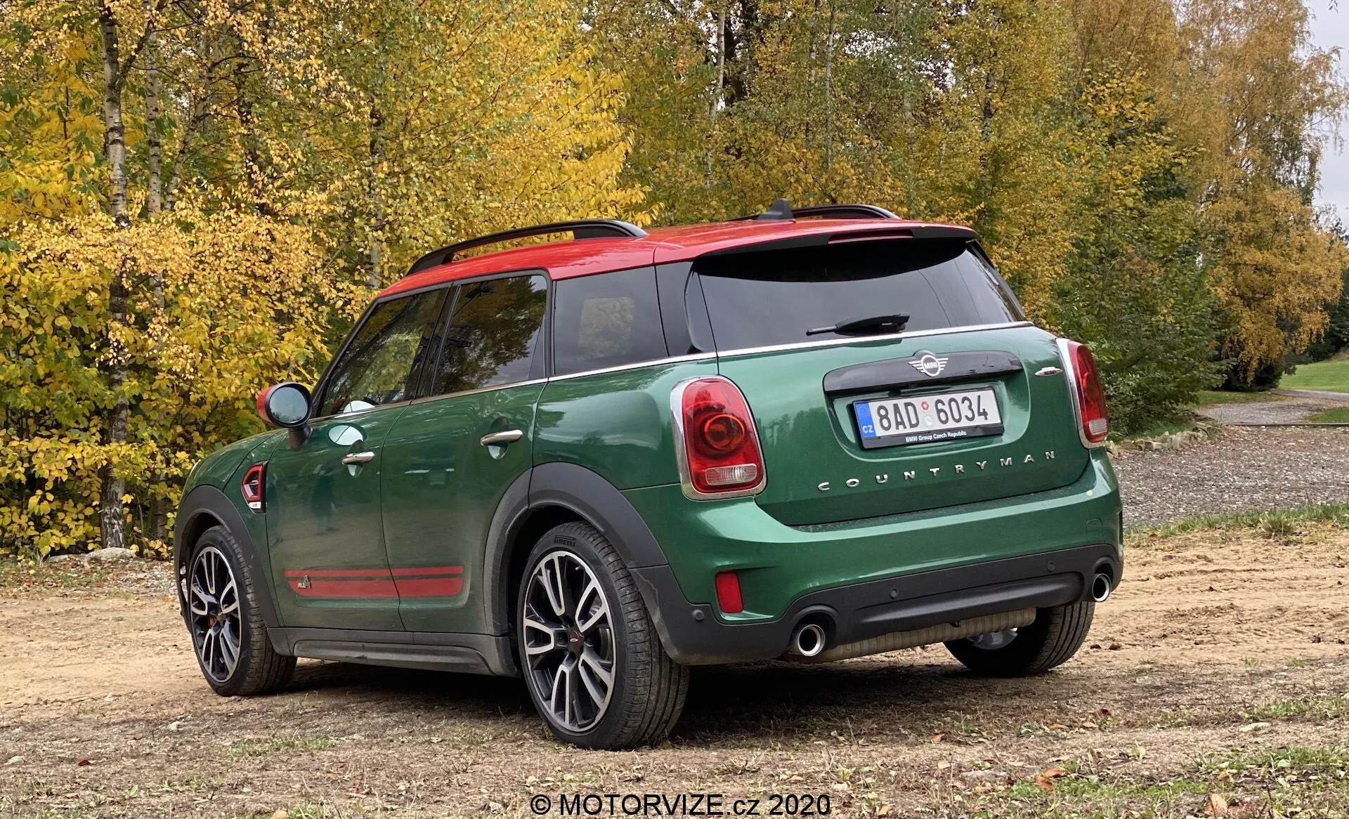 Widok z tyłu w trzech czwartych na Mini Cooper Countryman John Cooper Works 2020 z dwukolorowym ciemnozielonym i czarnym lakierem, czerwonymi kapturkami lusterek, widocznym emblematem Mini i napisami 'Countryman', wyjątkowymi tylne światła, wyraźny tylny zderzak z czujnikami parkowania, tylna szyba z odmrażaczem, podwójnymi rurami wydechowymi, tylną wycieraczką, pięciokołowymi felgami aluminiowymi, zaparkowany na polnej drodze z drzewami i jesiennymi liśćmi w tle, uchwycony pod nieznacznym wysokim kątem.