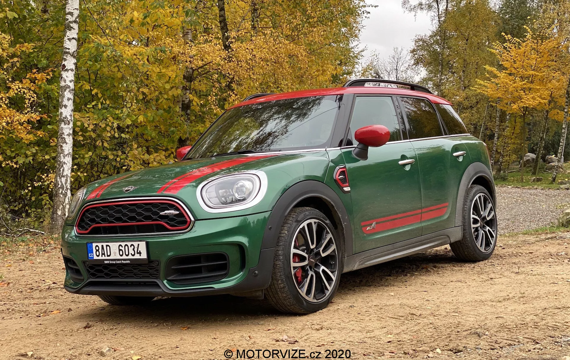 Widok z trzech czwartych przodu na Mini Countryman John Cooper Works z 2020 roku, ukazujący sportowy design z ciemnozielonym dwukolorowym nadwoziem i czerwonymi akcentami na lusterkach bocznych, kratce przedniej i progach bocznych. Cechy obejmują charakterystyczny wlot na masce, okrągłe reflektory, przednią kratkę z wzorem plastra miodu, duże sportowe felgi wieloramienne, czerwone zaciski hamulcowe, wyraźne progi boczne i agresywną sylwetkę. Samochód jest ustawiony na nieutwardzonej nawierzchni z leśnym otoczeniem w tle, pod naturalnym oświetleniem w pochmurny dzień.