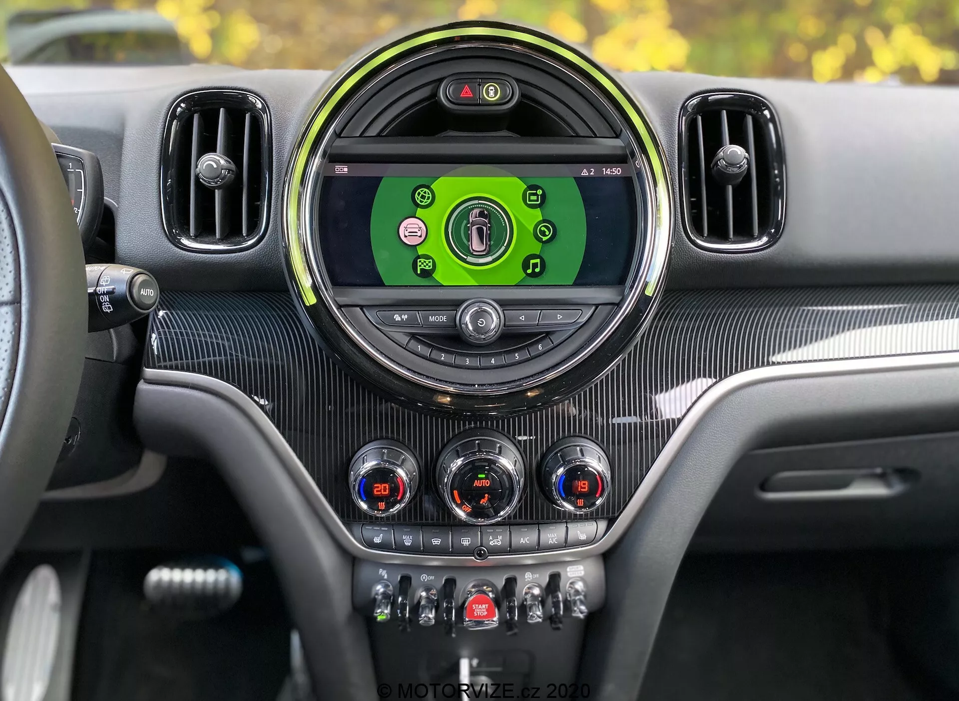 Deska rozdzielcza wewnątrz Mini Countryman John Cooper Works 2020 z perspektywy kierowcy. Konsola centralna z dużym, okrągłym wyświetlaczem infotainment pokazującym kolorowe ikony dla mediów, łączności telefonicznej, nawigacji i ustawień pojazdu. Otoczony stylową okrągłą ramką i flankowany przez dodatkowe kratki nawiewu. Poniżej wyświetlacza znajdują się analogowe pokrętła z chromowanymi akcentami do sterowania klimatyzacją i przełączniki do funkcji takich jak podgrzewanie siedzeń, odszranianie, recyrkulacja powietrza i tryby jazdy. Tekstura przypominająca włókno węglowe i metaliczne akcenty zapewniają współczesny i luksusowy design wnętrza z widocznym cyfrowym zestawem wskaźników wskazującym, że pojazd jest włączony.