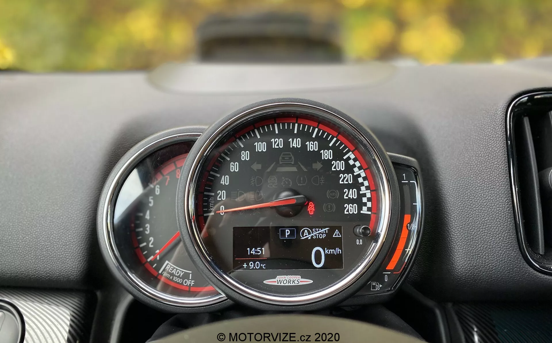 Szczegółowe zbliżenie na deskę rozdzielczą Mini Countryman John Cooper Works 2020. Skupienie na okrągłym prędkościomierzu o zakresie od 0 do 260 km/h, ze wskazówką na 0 km/h, co wskazuje, że samochód jest nieruchomy. Cyfrowy wyświetlacz na dole pokazuje samochód w trybie 'P', temperaturę zewnętrzną 9,0°C, przebieg 1451 km i ikonę otwartego bagażnika. Słowa 'Auto Start/Stop' sugerują system oszczędzania paliwa. Prędkościomierz jest otoczony chromowym pierścieniem i ma czerwoną strefę zaczynającą się tuż za 220 km/h. Po lewej stronie widać częściowy widok innego wskaźnika, prawdopodobnie obrotomierza. Deska rozdzielcza ma teksturowaną powierzchnię, a niewielki wlot powietrza jest częściowo widoczny po prawej stronie prędkościomierza.