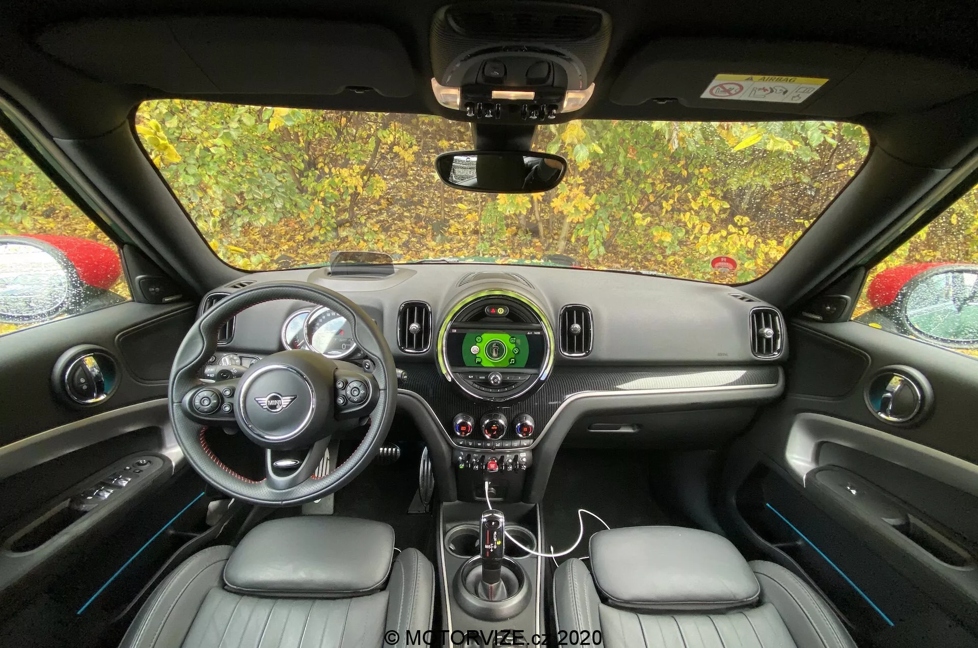 Wnętrze Mini Countryman 2020 John Cooper Works widziane z perspektywy tylnego siedzenia, skupiające się na kokpicie kierowcy i strefie kontroli. Widoczna jest kierownica ze srebrnymi akcentami i logo marki, centralny prędkościomierz z wyświetlaczem cyfrowym, elegancki design kokpitu, dźwignia zmiany biegów, sterowanie klimatem, fotele obszyte skórą oraz lusterko wsteczne.
