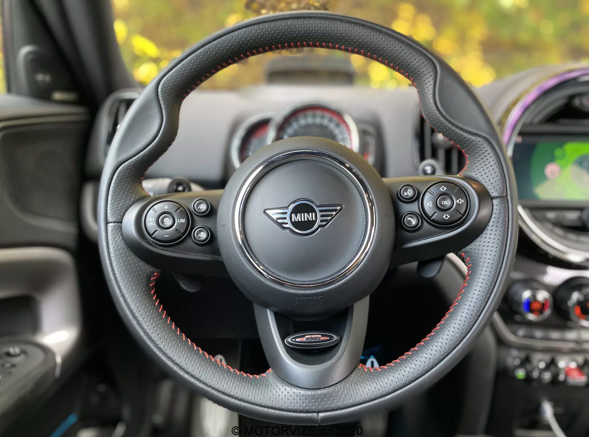 Widok wnętrza Mini Countryman John Cooper Works 2020 z perspektywy siedzenia kierowcy, skupiający się na kierownicy z logo Mini pośrodku. Trójramienna kierownica, pokryta skórą z czerwonymi szwami, posiada wielofunkcyjne kontrolki do zarządzania dźwiękiem, telefonem, kontrolą głosową i tempomatem. Widoczne są łopatki zmiany biegów, co sugeruje automatyczną skrzynię biegów z manualnym wyborem biegów. W tle znajduje się deska rozdzielcza z okrągłymi elementami, centralnym wyświetlaczem pokazującym mapę, wentylacją i kolorowymi akcentami.