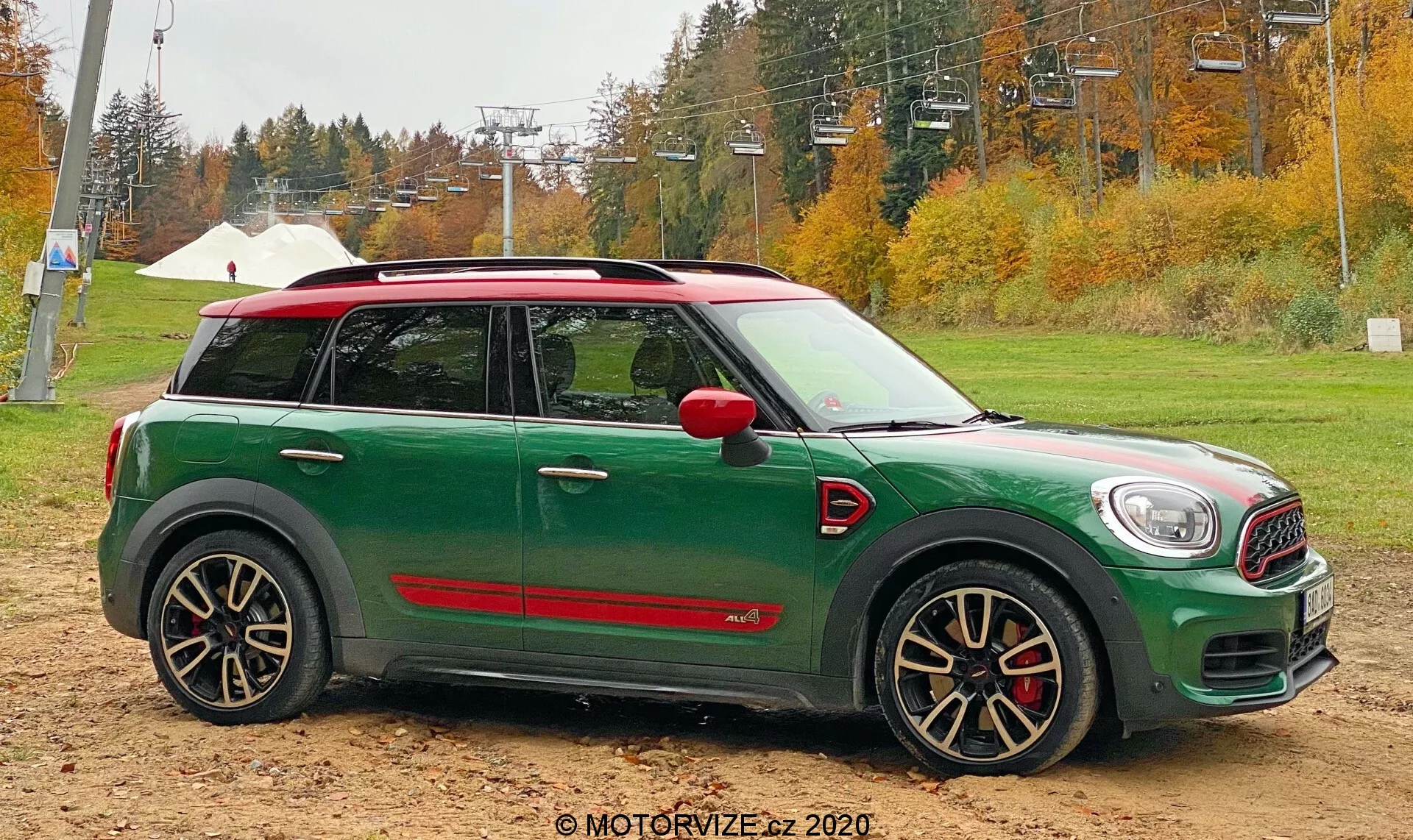 Boczny profil zielonego Mini Cooper Countryman 2020 John Cooper Works na piaszczystym podłożu z jesiennymi drzewami i infrastrukturą wyciągu narciarskiego w tle. Samochód ma biały dach, czerwone obudowy lusterek bocznych, agresywne czarno-srebrne felgi wieloramienne z czerwonymi zaciskami hamulcowymi oraz sportowe czerwone listwy boczne.