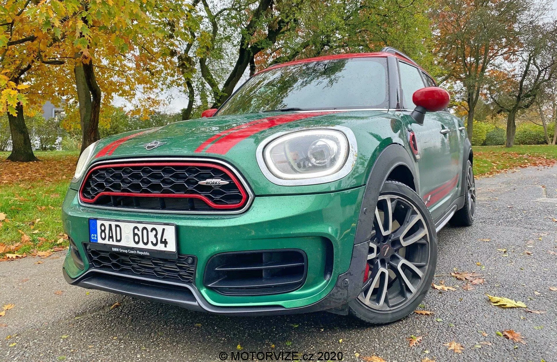 Obraz przedstawia hatchback Mini Countryman John Cooper Works 2020, uchwycony pod niskim kątem w przednim trzech czwartych. Widoczne są kluczowe elementy designu i technologii, w tym reflektory LED, charakterystyczna przednia krata, stylizowany zderzak, felgi aluminiowe, częściowo widoczne lusterka boczne z zintegrowanymi kierunkowskazami, aerodynamiczne linie nadwozia, odznaki i błyszczący lakier samochodu. Ta perspektywa podkreśla agresywną postawę i dynamiczny wygląd pojazdu.