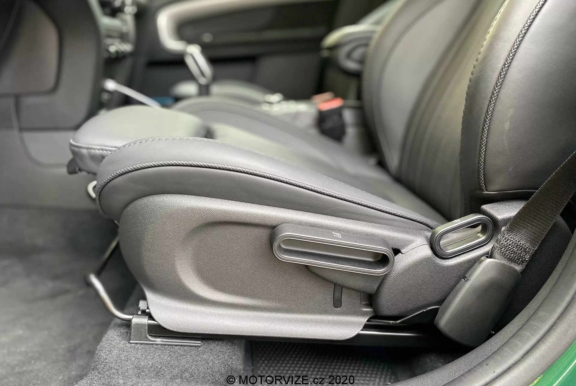 Zbliżenie na wnętrze Mini Countryman John Cooper Works 2020, podkreślając dolną boczną część siedzenia kierowcy z detalicznymi mechanizmami regulacji siedzenia. Siedzenie jest wyściełane ciemną skórą lub skórzanym materiałem, z wyraźnym bocznym wzmocnieniem, co wskazuje na komfort i jakość. Widoczne są manualne elementy regulacyjne, w tym uchwyt prętowy i okrągłe pokrętło, co sugeruje ergonomiczny design i regulację według preferencji użytkownika. Obecny jest także uchwyt pasa bezpieczeństwa, co świadczy o dbałości o bezpieczeństwo pasażerów.