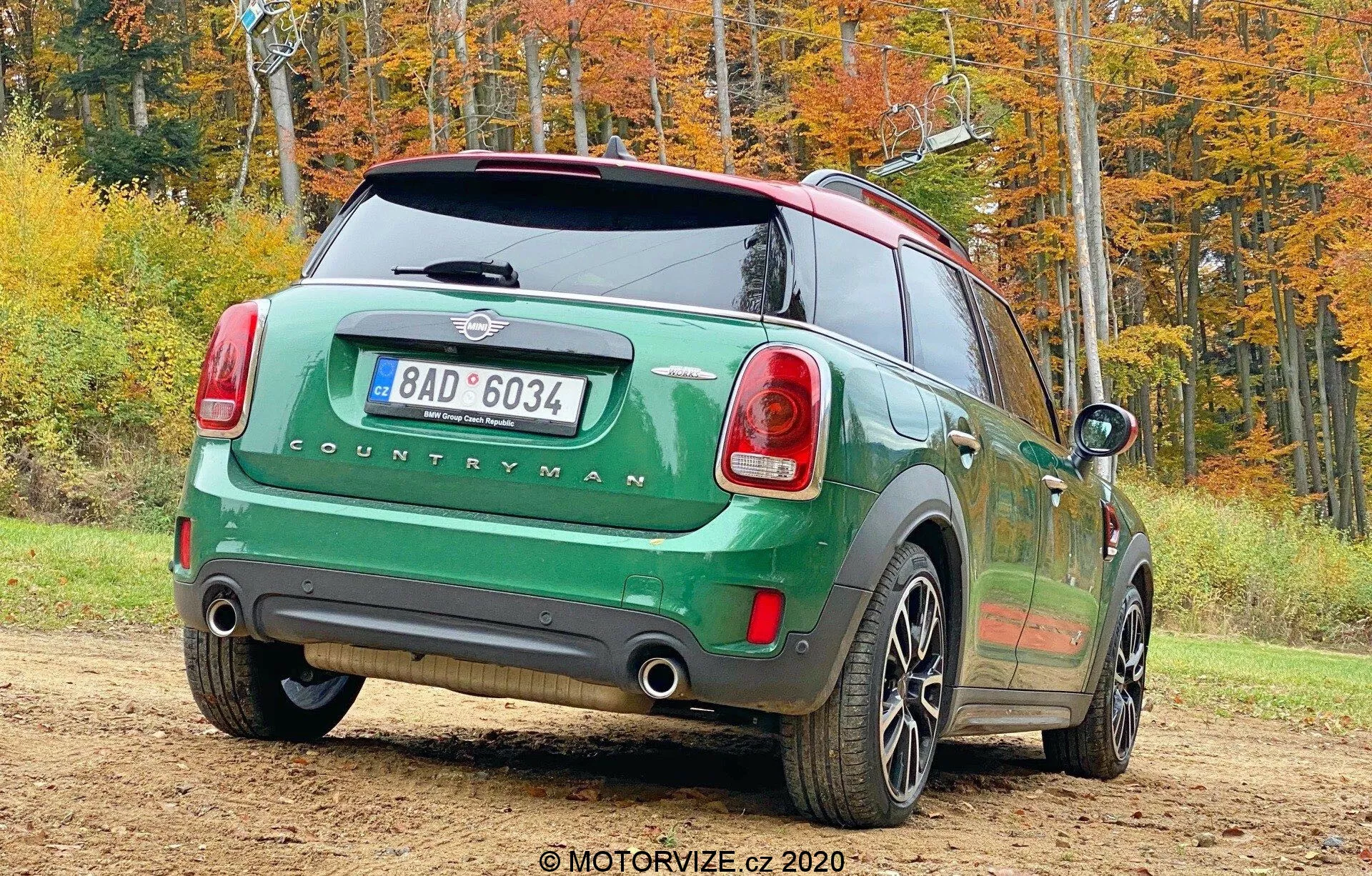 Obraz zielonego Mini Countryman 2020 modelu John Cooper Works zaparkowanego na zewnątrz w jesiennym otoczeniu, zrobiony pod kątem trzech czwartych od tylnej strony. Prawa tylna strona samochodu jest skierowana w stronę kamery i pokazuje tylne światła, tylną szybę, znak 'COUNTRYMAN', podwójny układ wydechowy, pięcioramienne koła, kontrastowy czarny dach i lusterka boczne. Obraz zawiera również tylna tablicę rejestracyjną, tylna wycieraczkę i antenę do łączności.