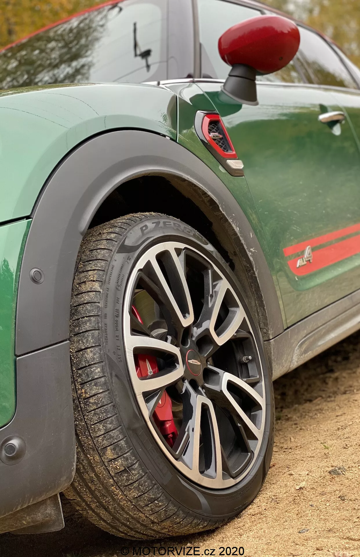 Zbliżenie tylnego koła i części tylnego błotnika Mini Countryman John Cooper Works 2020. Zdjęcie wykonane jest z boku pod nieco ukośnym, niskim kątem, aby podkreślić wieloszprychowe aluminiowe koło w szarym lub srebrnym wykończeniu. Czerwony zacisk hamulcowy sugeruje wysokowydajny system hamulcowy. Opona ma widoczne bieżniki, co oznacza, że jest odpowiednia do różnych warunków drogowych. Zielony samochód ma stylizowany pasek wzdłuż dolnej części drzwi oraz częściowo widoczny emblemat, co dodaje sportowego wyglądu. Widoczny jest również lusterko boczne z czerwonym kapturem i chlapacz, co zwiększa dynamiczny wygląd pojazdu. Rozmyte tło koncentruje uwagę na tych detalach, idealnie podkreślając elementy wydajnościowe samochodów.