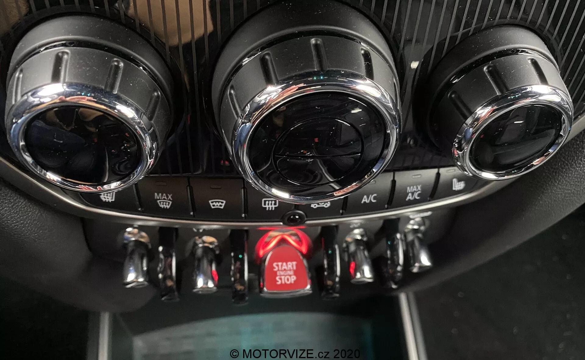 To zdjęcie przedstawia zbliżenie konsoli środkowej deski rozdzielczej w Mini Countryman John Cooper Works z 2020 roku, z naciskiem na system kontroli klimatu i przycisk start-stop silnika. Konsola pokazuje dwa okrągłe otwory wentylacyjne z metalicznymi wykończeniami u góry, szczegółowy panel sterowania klimatyzacją z trzema metalicznymi pokrętłami do regulacji temperatury, prędkości wentylatora i kierunku przepływu powietrza oraz kilkoma przyciskami do funkcji takich jak odmrażanie i recyrkulacja powietrza. Poniżej znajduje się czerwony, łatwo zauważalny przycisk do uruchamiania i wyłączania silnika. Tekstury panelu obejmują sportowy design przypominający włókno węglowe wokół otworów wentylacyjnych oraz standardowy czarny plastik dla dolnego panelu. Kąt zdjęcia, z przodu i nieco powyżej, podkreśla wysokiej jakości design i zaawansowane funkcje.
