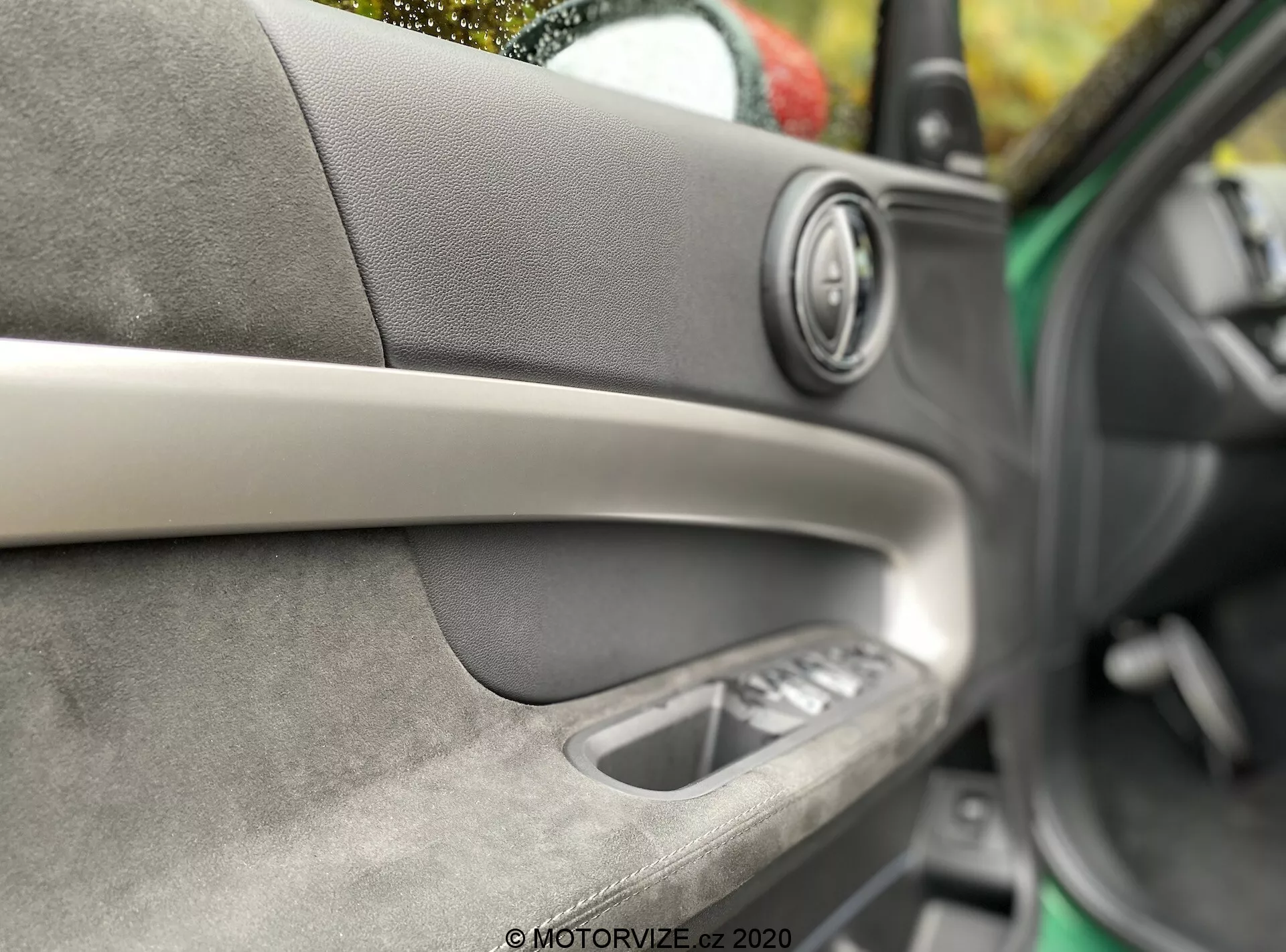 Szczegółowy widok wnętrza drzwi samochodu Mini Countryman John Cooper Works 2020. Skupiony na głośniku audio premium z metalicznym pierścieniem, ukośnym pasku dekoracyjnym, miękkim materiale z widocznymi szwami, wewnętrznej klamce drzwi oraz sterowaniu szybami elektrycznymi. Mała głębia ostrości podkreślająca rzemiosło i komfort wnętrza.