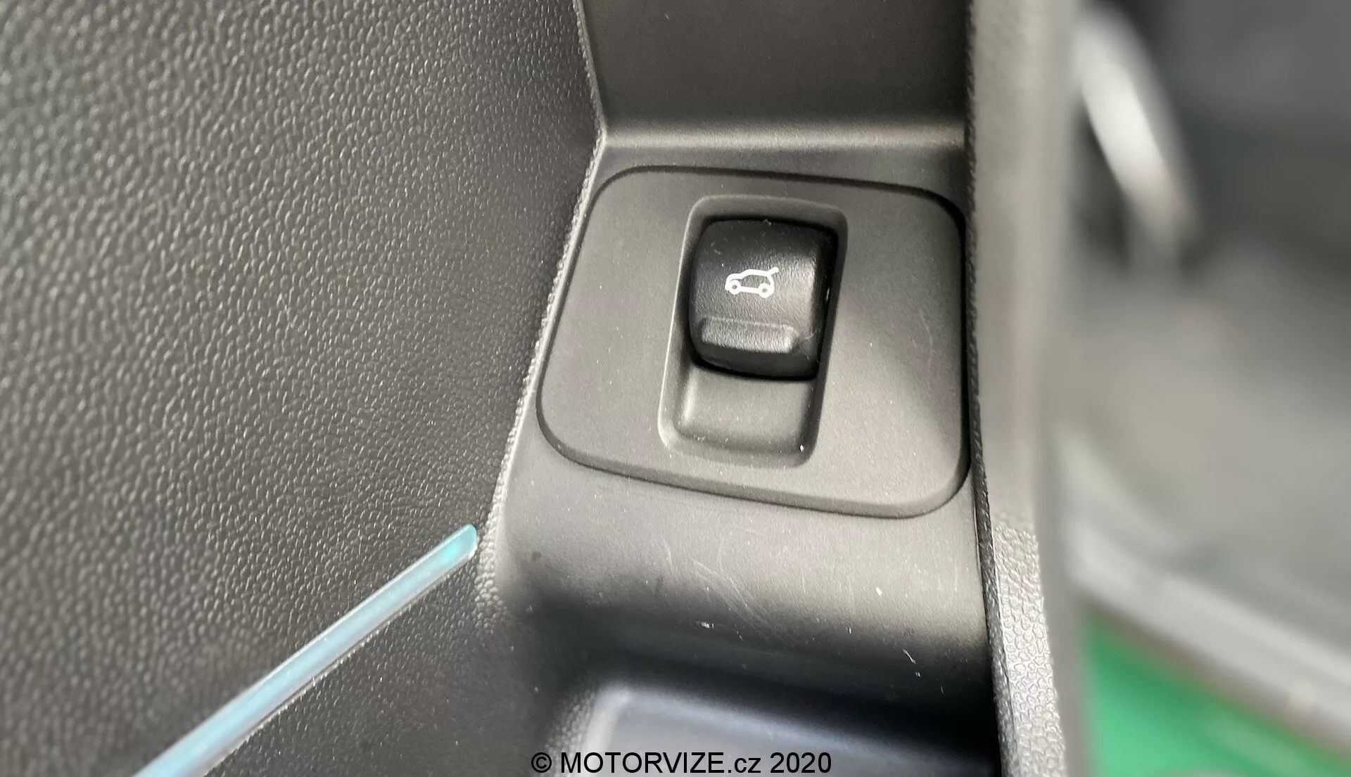 Zbliżenie na przełącznik sterowania lusterkami bocznymi wewnątrz Mini Countryman 2020 John Cooper Works. Prostokątny przełącznik z zaokrąglonymi narożnikami, umieszczony na płaskim panelu, jest wyróżniony symbolem lusterka. Jedna strona przełącznika jest w cieniu, co sugeruje źródło światła po przeciwnej stronie. Tło to teksturowany czarny plastik i cienka niebieska linia oświetlenia ambientowego, wzbogacając estetykę wnętrza pojazdu.