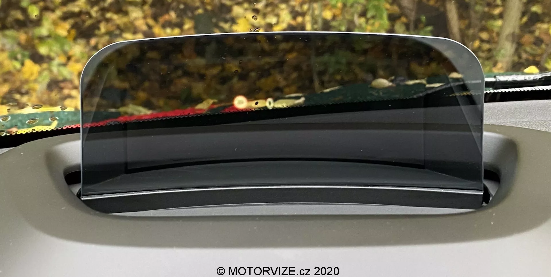 Obrazek przedstawiający wyświetlacz head-up (HUD) w Mini Countryman 2020, wyświetlany na przezroczystym ekranie nad deską rozdzielczą z perspektywy kierowcy. Przezroczysty, szklany ekran HUD jest płynnie zintegrowany z wnętrzem samochodu, zaokrąglony i lekko nachylony dla optymalnej widoczności. Tło za przednią szybą jest rozmyte, co podkreśla skupienie na HUD, który prezentuje kluczowe informacje dla kierowcy.