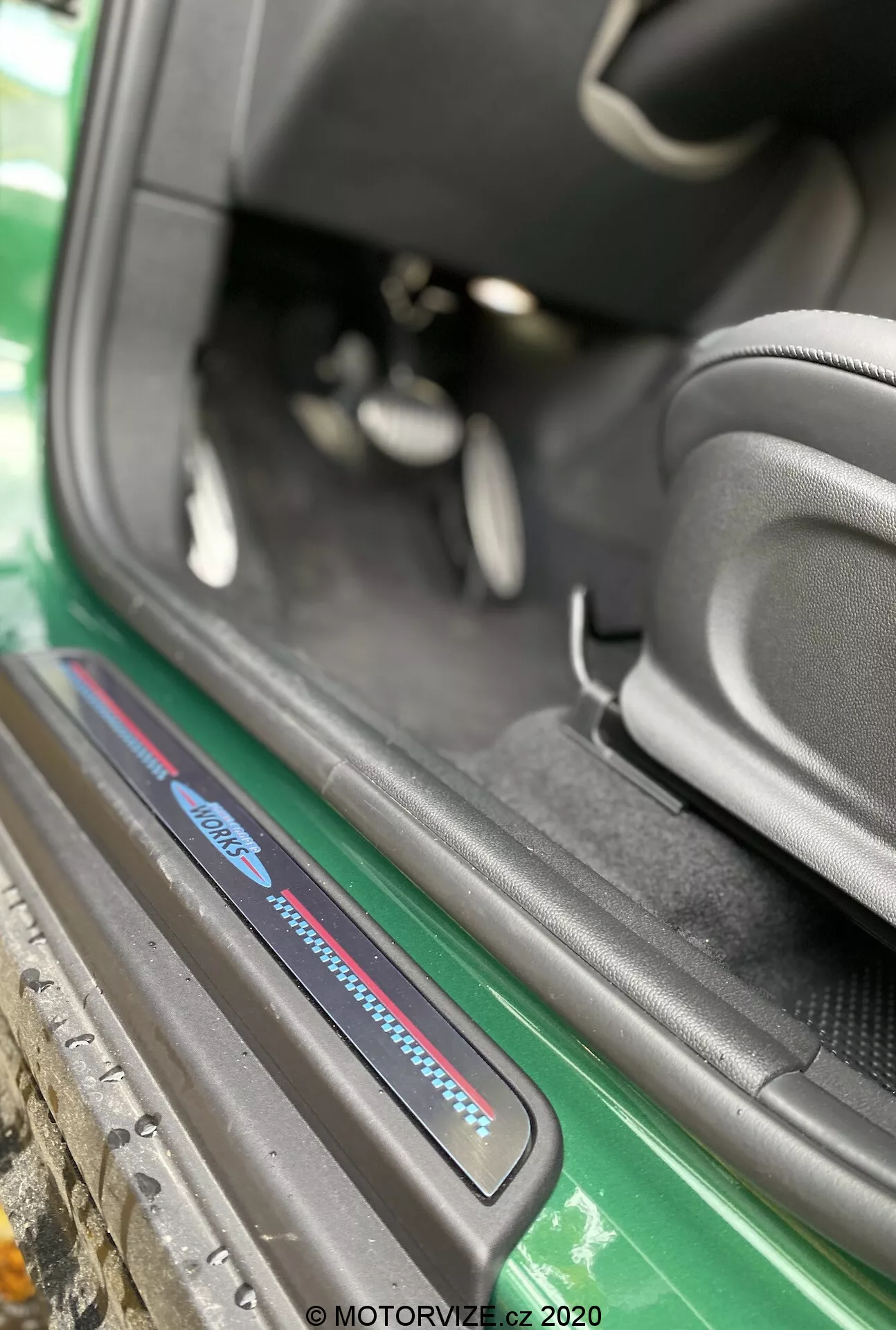 Zbliżenie na obszar progu drzwi Mini Countryman John Cooper Works 2020 z ukośnego kąta skupionego na progu po stronie pasażera. Obraz, uchwycony z zewnątrz pojazdu, podkreśla dolną część wnętrza samochodu, w tym płytkę progu z niebiesko-czerwonymi akcentami, czarne dywaniki, sportowy boczny podparcie czarnego fotela, częściowy widok pedałów i dobrze utrzymany obszar progu. Widoczne elementy zewnętrzne obejmują zieloną spódnicę boczną i nadwozie, podkreślając atrakcyjność estetyczną. Znak wodny 'MOTORVIZE.cz 2020' sugeruje, że zdjęcie zostało wykonane w 2020 roku na potrzeby recenzji motoryzacyjnej.
