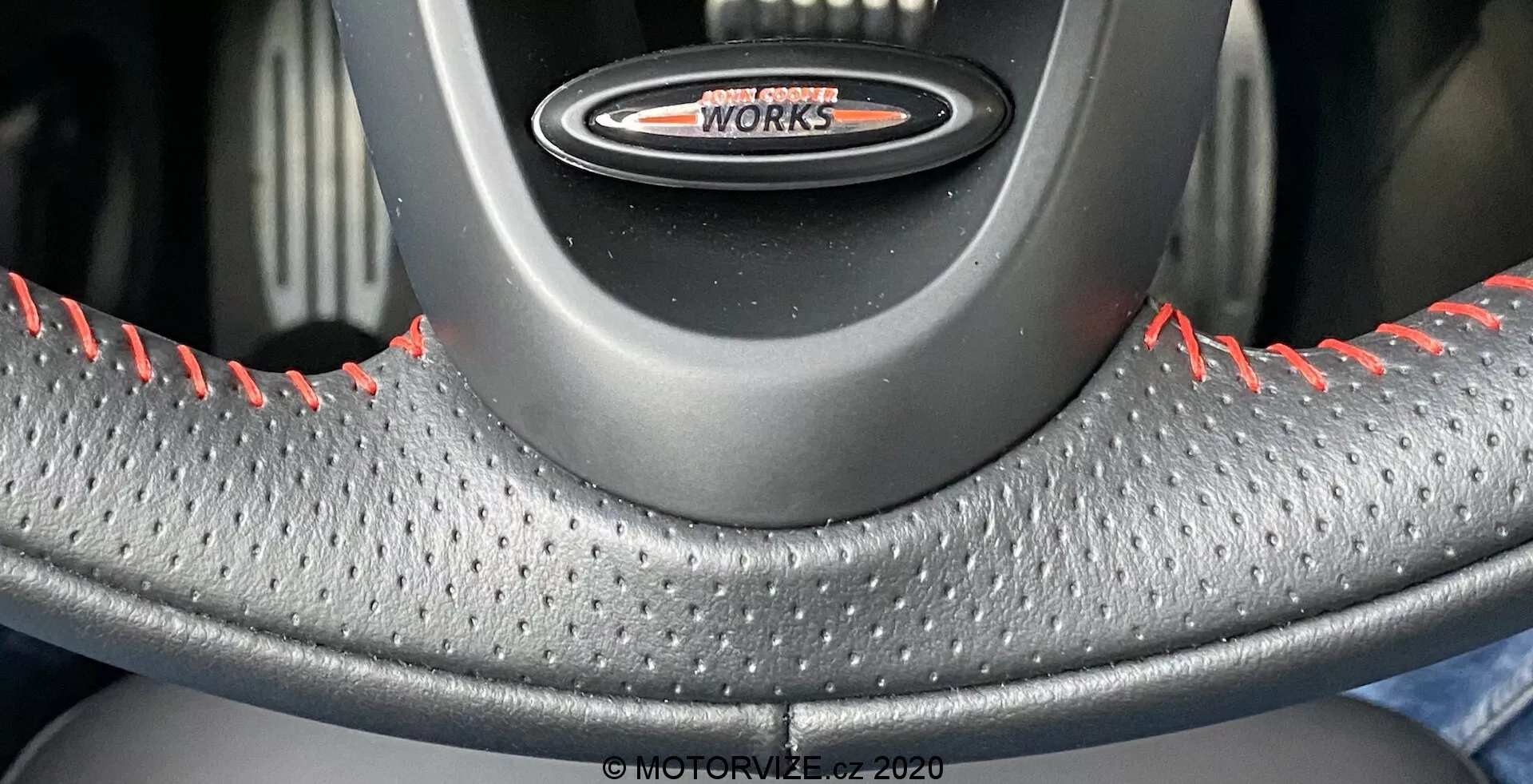 Zbliżenie na kierownicę z perspektywy siedzenia kierowcy, z emblematem 'John Cooper Works', skórzanym pokryciem z teksturą wgłębień dla lepszego chwytu i kontrastującymi czerwonymi przeszyciami wskazującymi na wysokowydajne warianty MINI Cooper