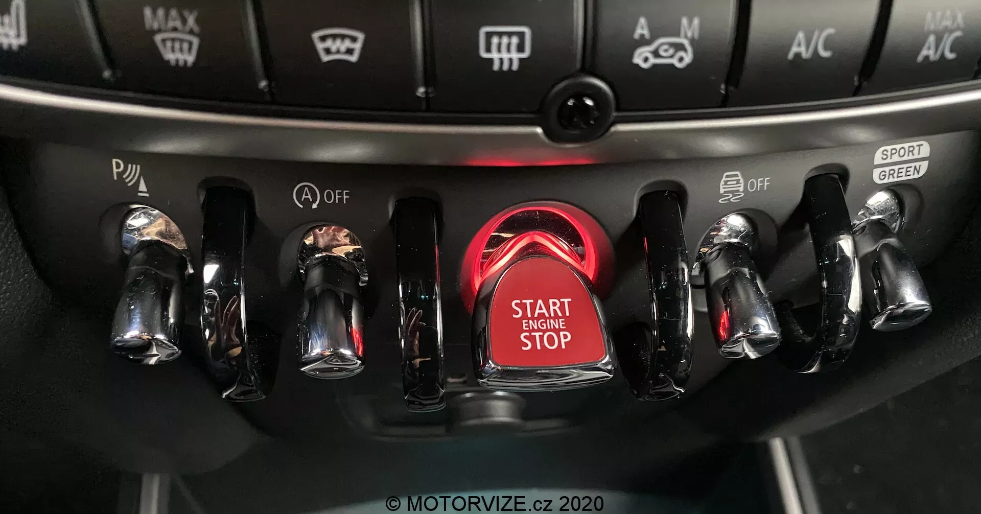 Zbliżenie środka konsoli Mini Countryman John Cooper Works 2020 z naciskiem na obszar zapłonu, podkreślające przycisk uruchamiania/zatrzymywania silnika otoczony świecącym czerwonym pierścieniem, z metalicznymi przełącznikami wykończonymi chromem po obu stronach do różnych sterowników pojazdu, w tym trybów jazdy i ustawień klimatyzacji.