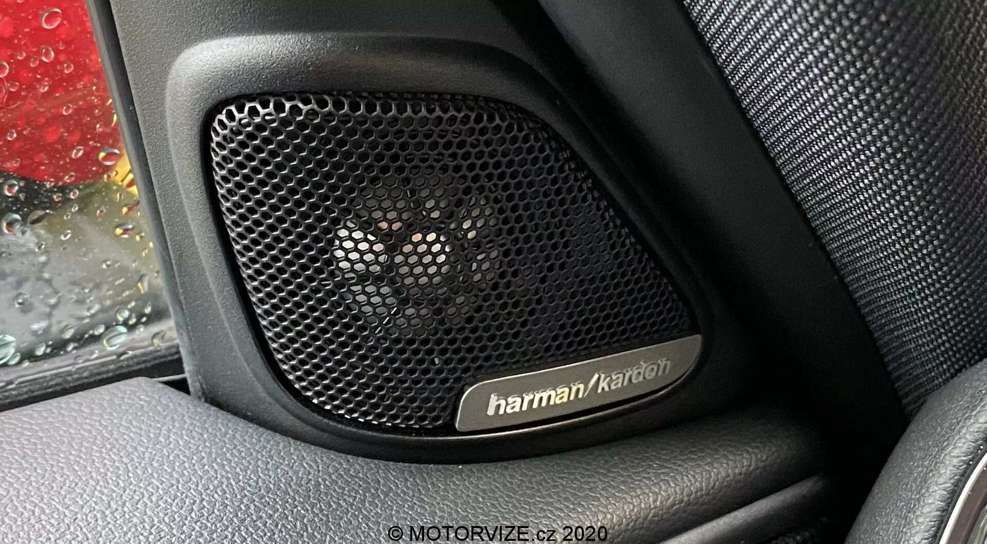 Zbliżenie na system głośników Harman Kardon wewnątrz panelu drzwi Mini Countryman 2020 John Cooper Works. Fotografia skupia się na perforowanej kratce głośnika, zaprojektowanej w celu zapewnienia przezroczystości akustycznej i ochrony, osadzonej na teksturowanym wnętrzu wyglądającym jak skóra. Kropelki deszczu są widoczne na szybie samochodu, dodając kontekstu zdjęciu. System audio premium podkreśla kluczowe funkcje technologiczne, ukazując luksus i trwałość projektu pojazdu.