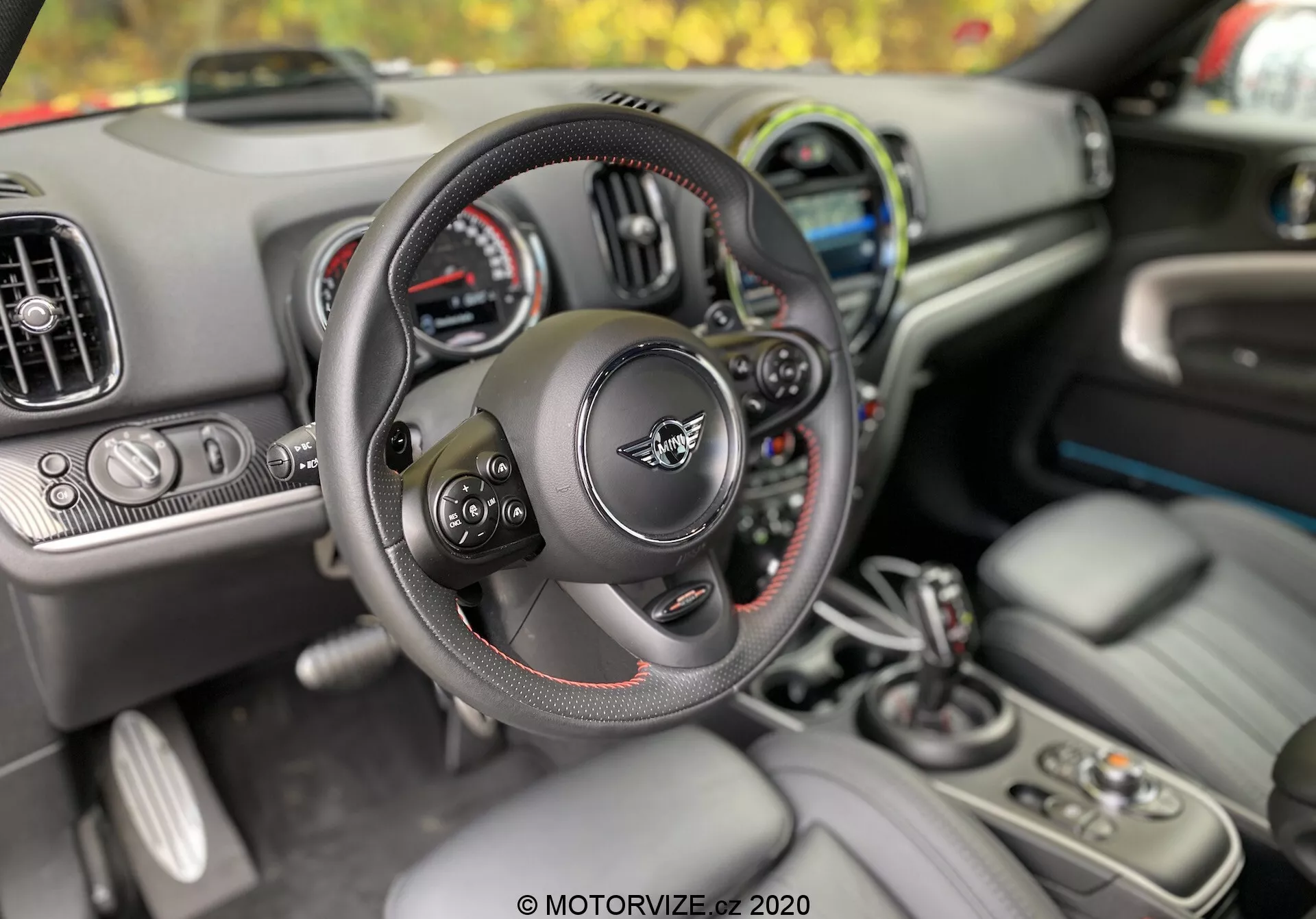 Widok wnętrza Mini Countryman 2020 z perspektywy kierowcy, podkreślający wielofunkcyjną kierownicę z czerwoną nicią, zestaw wskaźników z analogowymi i cyfrowymi wyświetlaczami, otwory wentylacyjne i ekran infotainment. Na konsoli środkowej widoczne są dźwignia zmiany biegów, uchwyty na kubki, przyciski sterujące i brelok wskazujący na system bezkluczykowy i rozrusznik z przyciskiem. Przedni plan skupia się na kierownicy i dźwigni zmiany biegów, ukazując ergonomię i komfort zaprojektowane dla kierowcy.