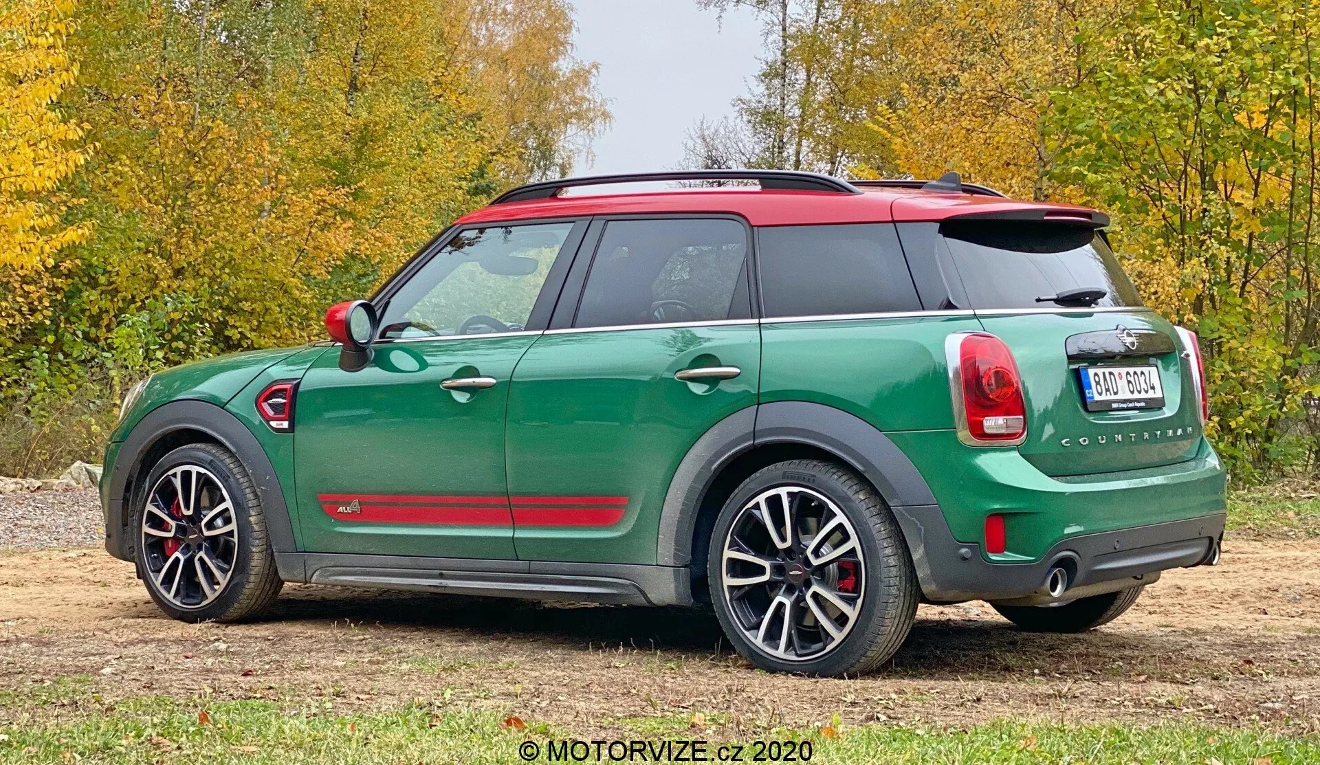 Obraz przedstawia widok od trzech czwartych tyłu na zielone Mini Cooper Countryman z 2020 roku z kontrastowym czerwonym dachem i lusterkami. Cechy obejmują spoiler dachowy, relingi dachowe, tylne światła inspirowane Union Jack, tylna wycieraczka, podwójny wydech i czujniki parkowania. Pojazd jest wyposażony w duże aluminiowe koła i ochronę wokół nadkoli. Zdjęcie zostało zrobione na zewnątrz na żwirowej nawierzchni z trawą i liśćmi w tle.
