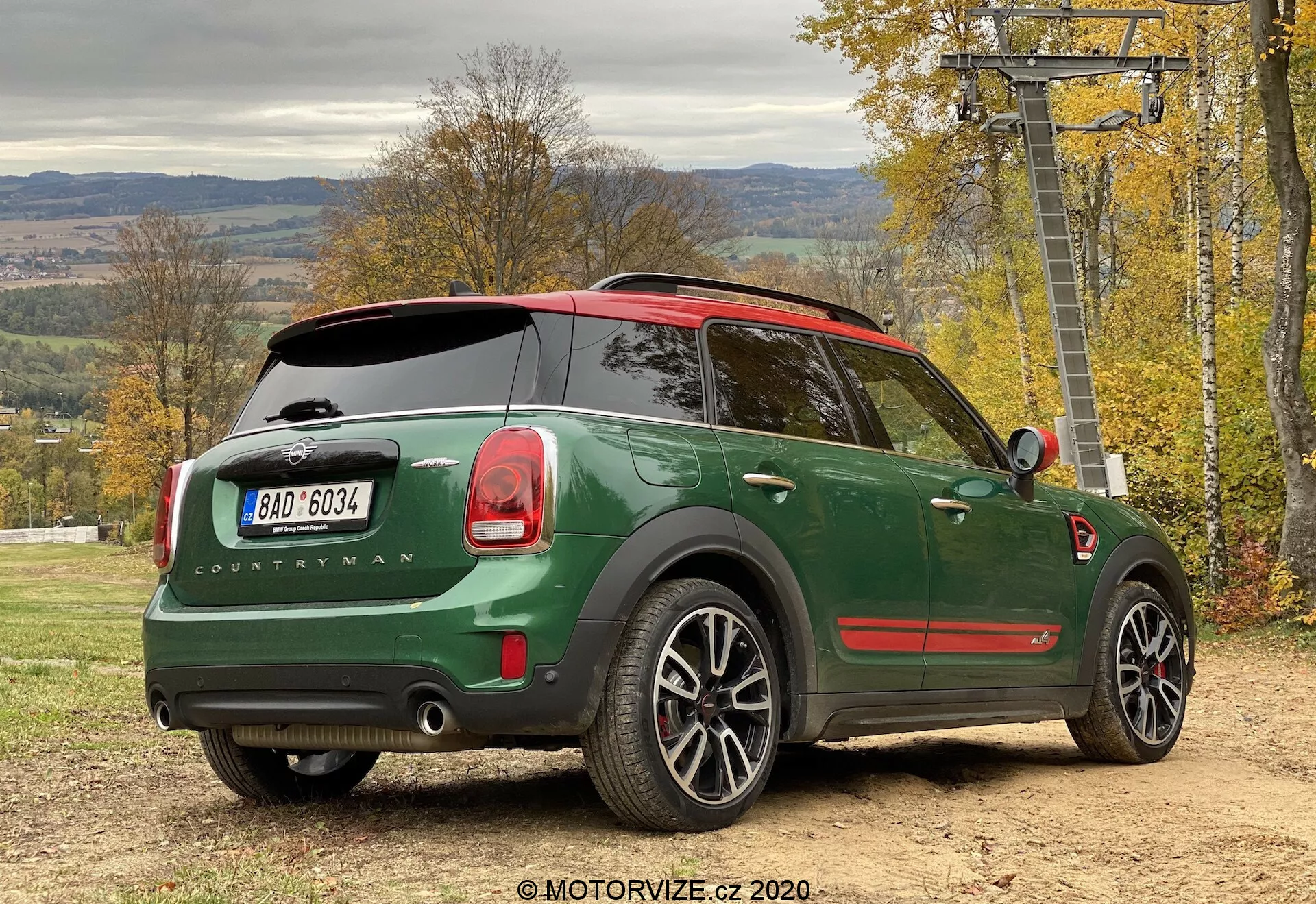 Widok zza tylnej trzy czwartej zielonego Mini Cooper Countryman John Cooper Works 2020 zaparkowanego na zewnątrz, podkreślający stylizację samochodu z perspektywy boczno-tylnej. Charakterystyczne cechy obejmują ciemnozielone nadwozie z czerwonym dachem, oznaczenie 'Countryman', logo Mini na tylnej klapie, charakterystyczne tylne światła, podwójny układ wydechowy, felgi aluminiowe, panoramiczny dach, wycieraczkę tylnej szyby, przyciemniane tylne szyby, czujniki parkowania oraz lusterko boczne z wbudowanym kierunkowskazem. Pojazd znajduje się na nieutwardzonej powierzchni z jesiennym listowiem i wyciągiem narciarskim w tle, sugerując aktywny styl życia na świeżym powietrzu.