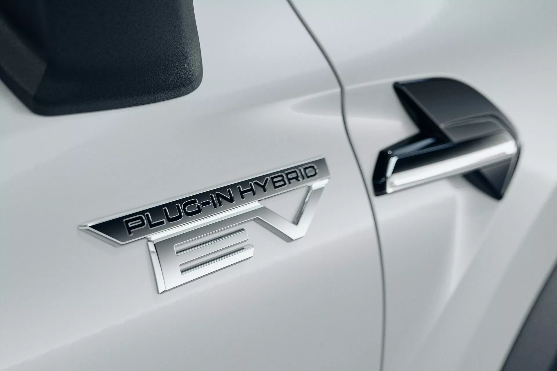 Zbliżenie na bok białego samochodu Mitsubishi ASX 2023 drugiej generacji, z wyróżniającym się emblematem 'PLUG-IN HYBRID EV', eleganckim czarnym lusterkiem bocznym z wbudowanym kierunkowskazem i błyszczącą, białą, aerodynamiczną karoserią.